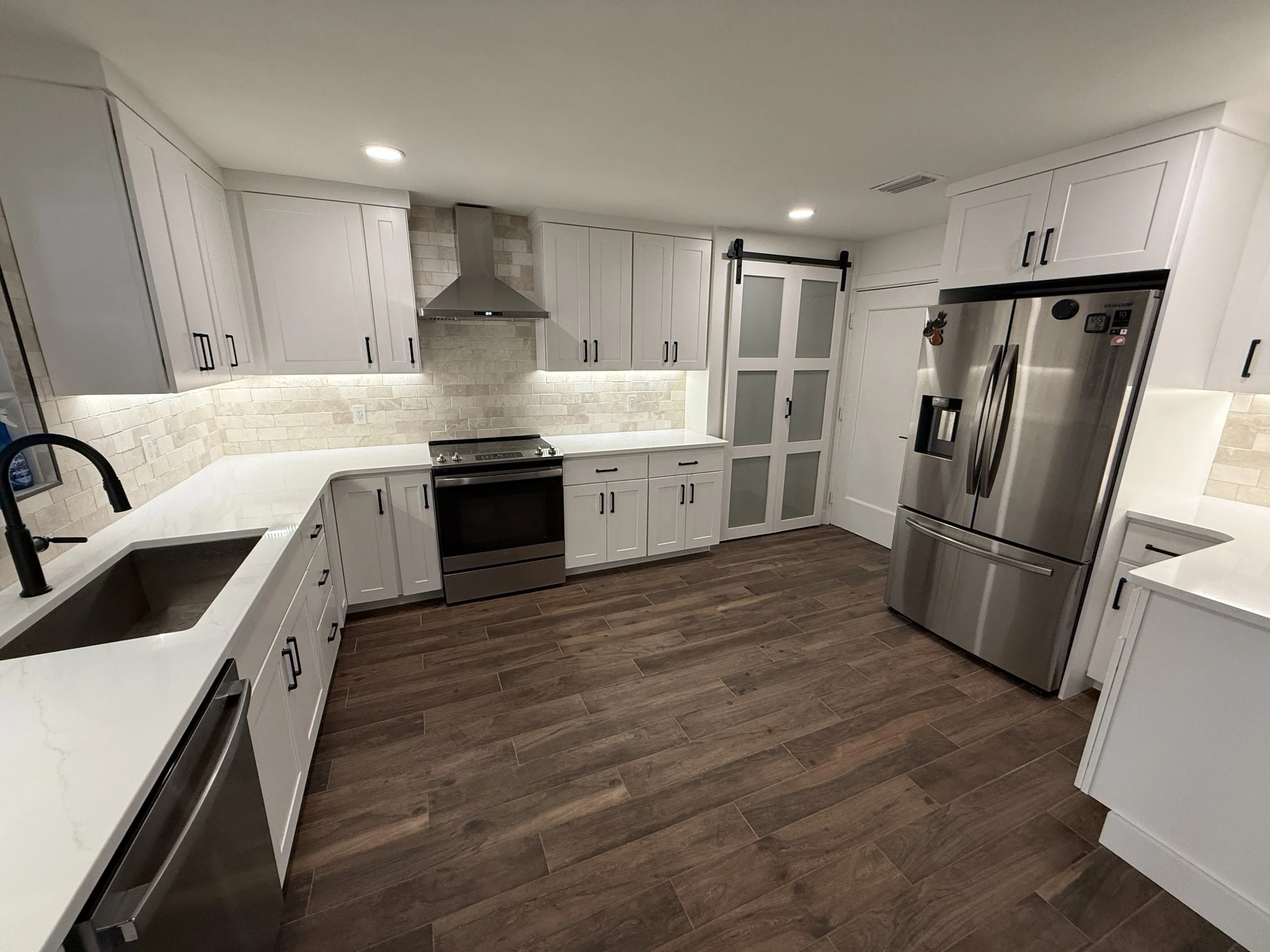 A modern kitchen with white cabinets, a stainless steel refrigerator, a black oven, and dark wood flooring. There is a grey brick backsplash, a white countertop with a black faucet, and a door with frosted glass and a black sliding barn door mechanism.