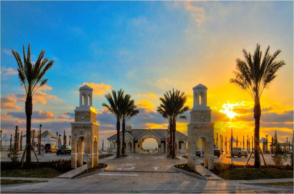 Entrance gate with two towers, palm trees, cars, and a beach in the background at sunset.