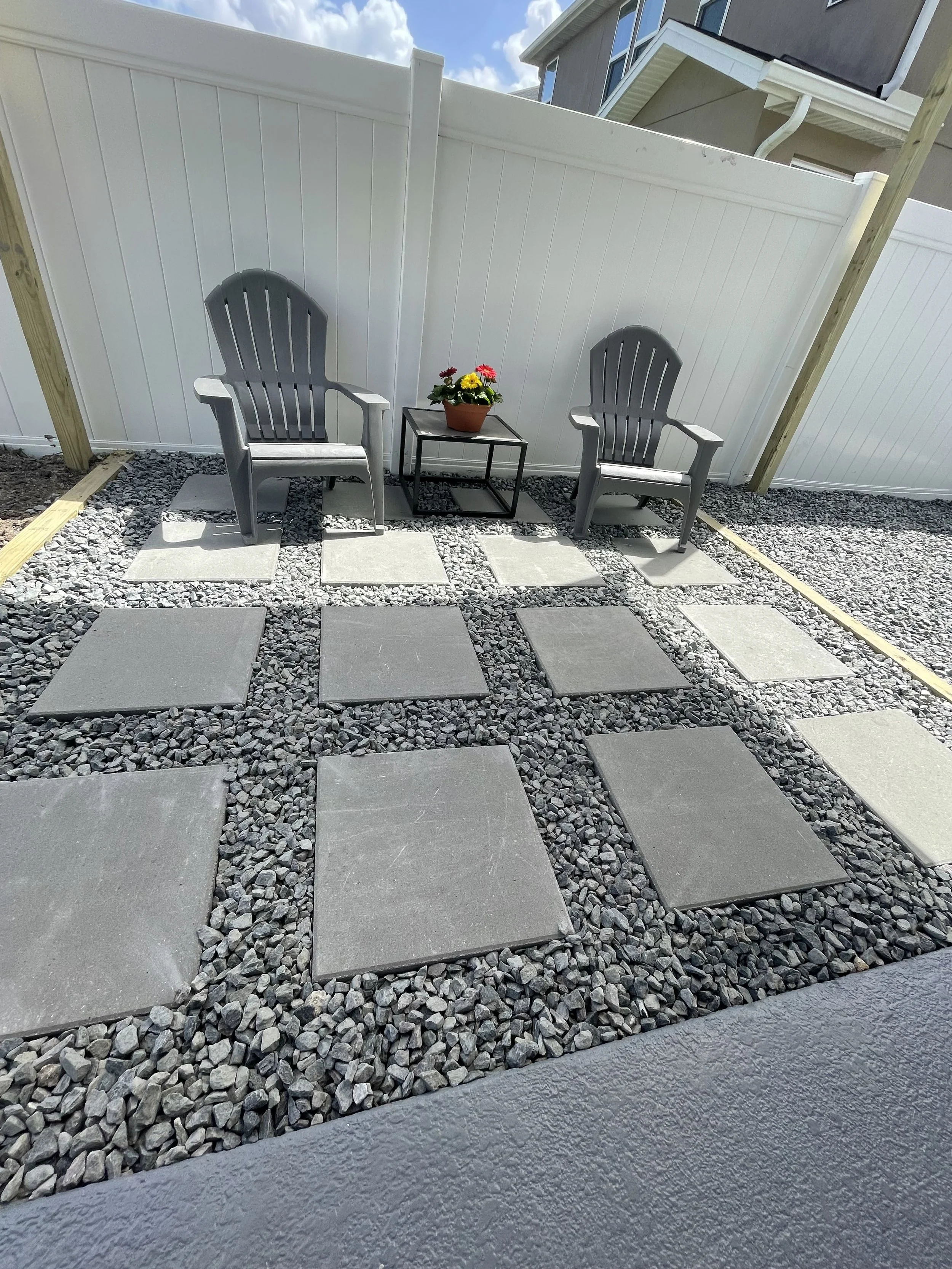 A backyard patio area with two gray Adirondack chairs and a small black metal table holding a potted flower arrangement, surrounded by square stepping stones and gray gravel, with a white privacy fence and a cloudy sky in the background.