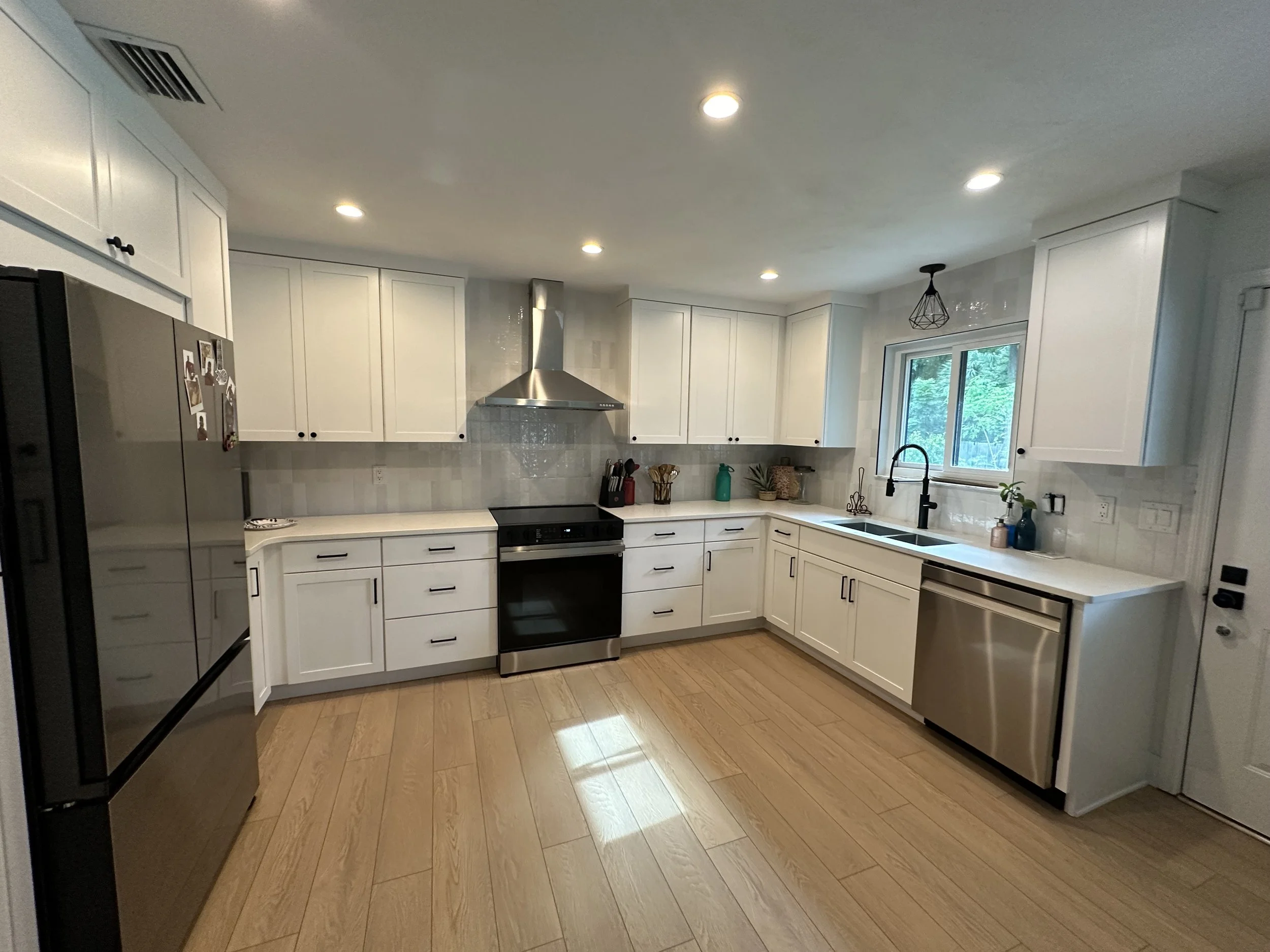 Modern white kitchen with wooden floors, stainless steel appliances, and a window with greenery outside.