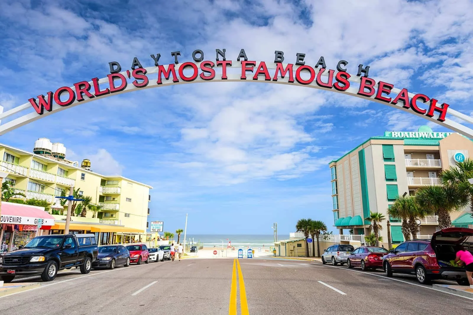 A street leading to a beach with an arch that reads 'Daytona Beach - World's Most Famous Beach'. There are cars parked along the sides, multi-story buildings, palm trees, and a view of the ocean in the background under a partly cloudy sky.
