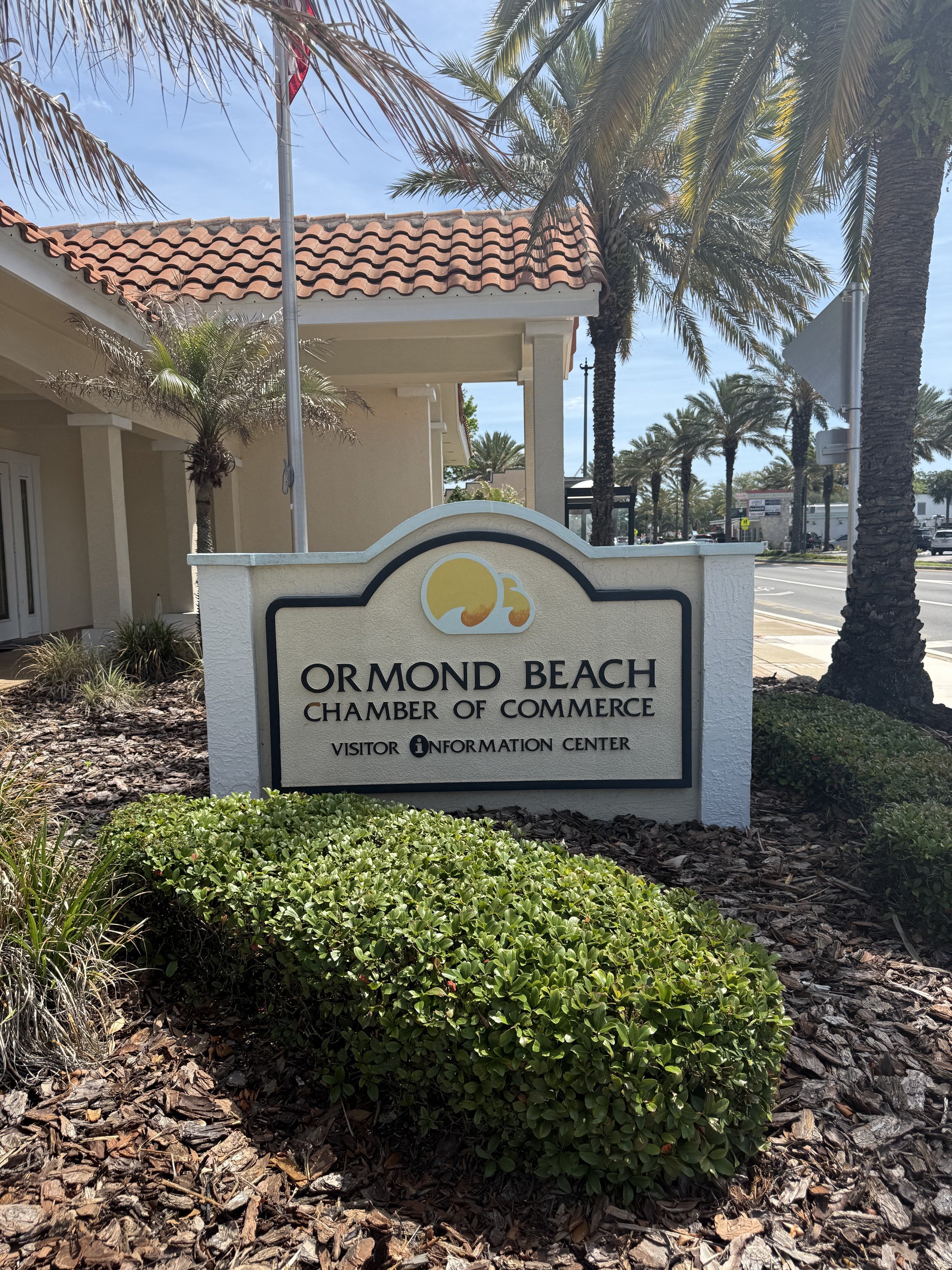 Sign for Ormond Beach Chamber of Commerce and Visitor Information Center with palm trees and building in the background.