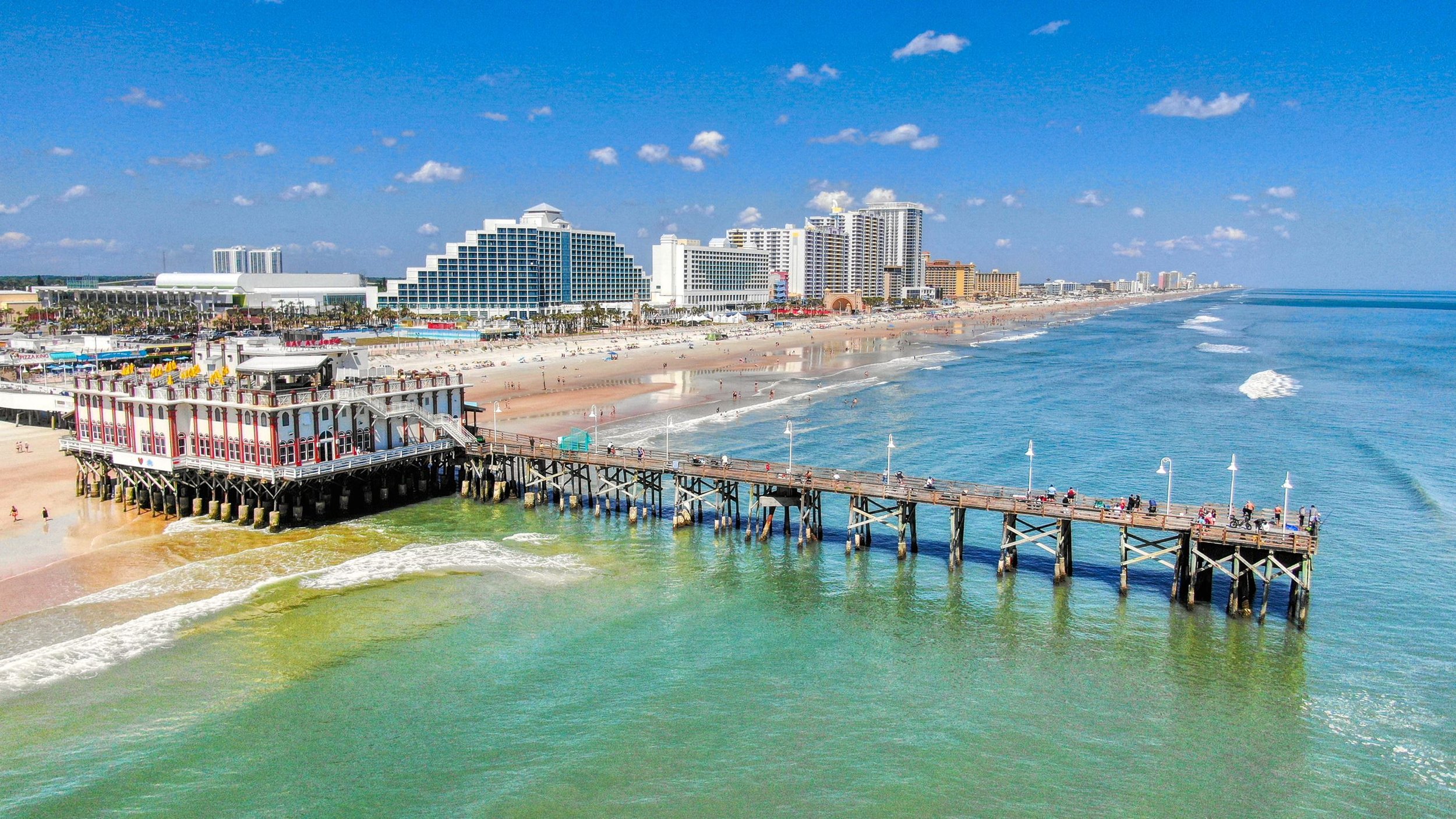 A sunny beach scene with a pier, colorful buildings, and high-rise hotels along the shoreline, with people walking on the beach and pier.