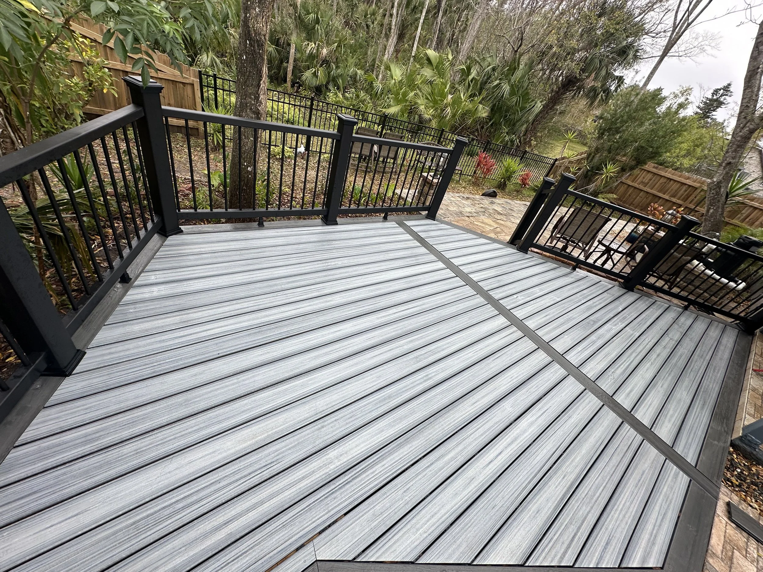 A backyard outdoor patio with a Trex composite deck and Trex Composite black railing, overlooking a garden with trees, shrubs, and a flagstone pathway. that we completed for a client in Ormond Beach Florida