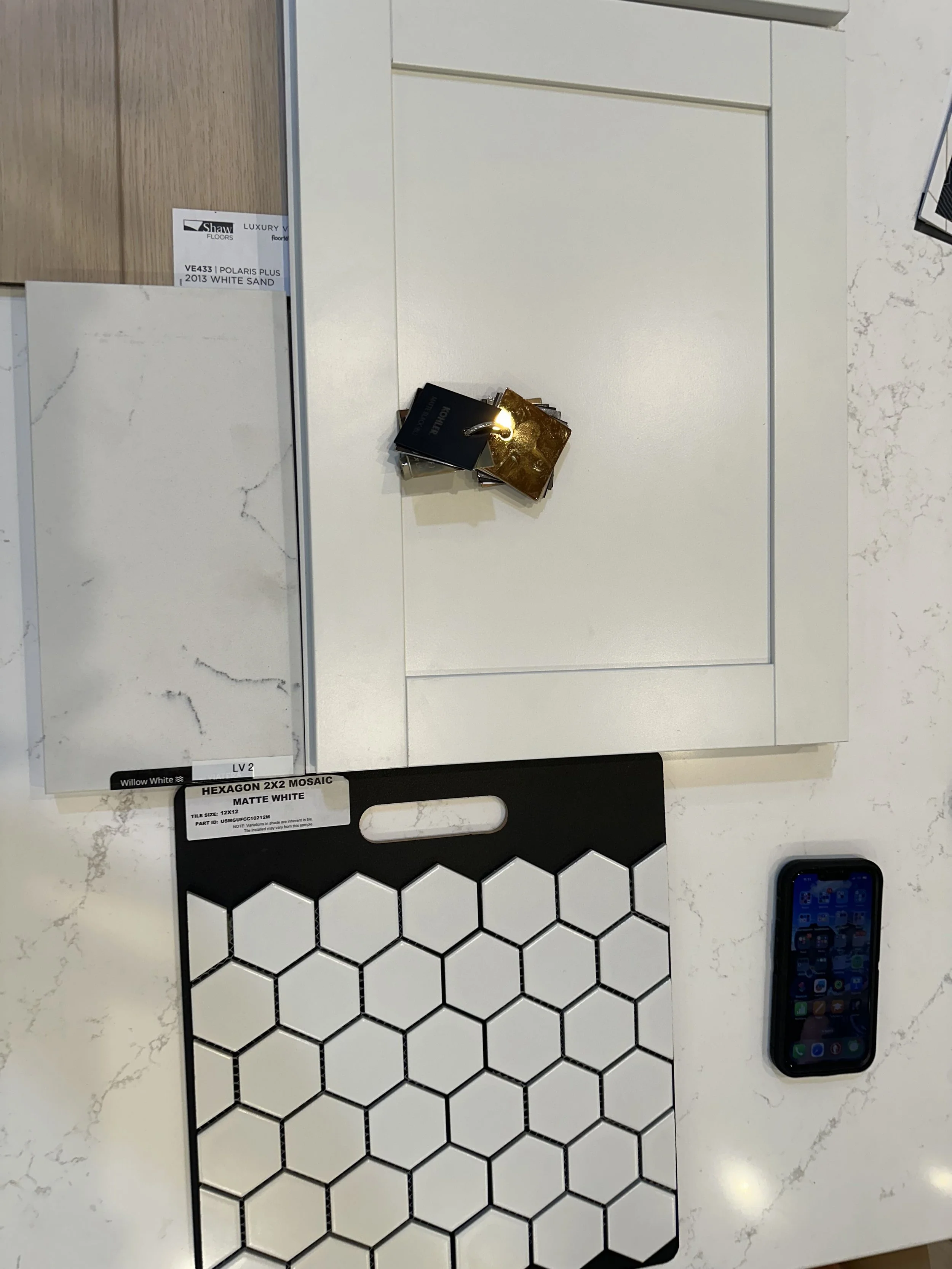 Display of tile options with a black and white hexagon mosaic tile at the bottom, sample wood flooring options, and small sample tiles on a white marble countertop. A smartphone with a colorful app grid is in the lower right corner.
