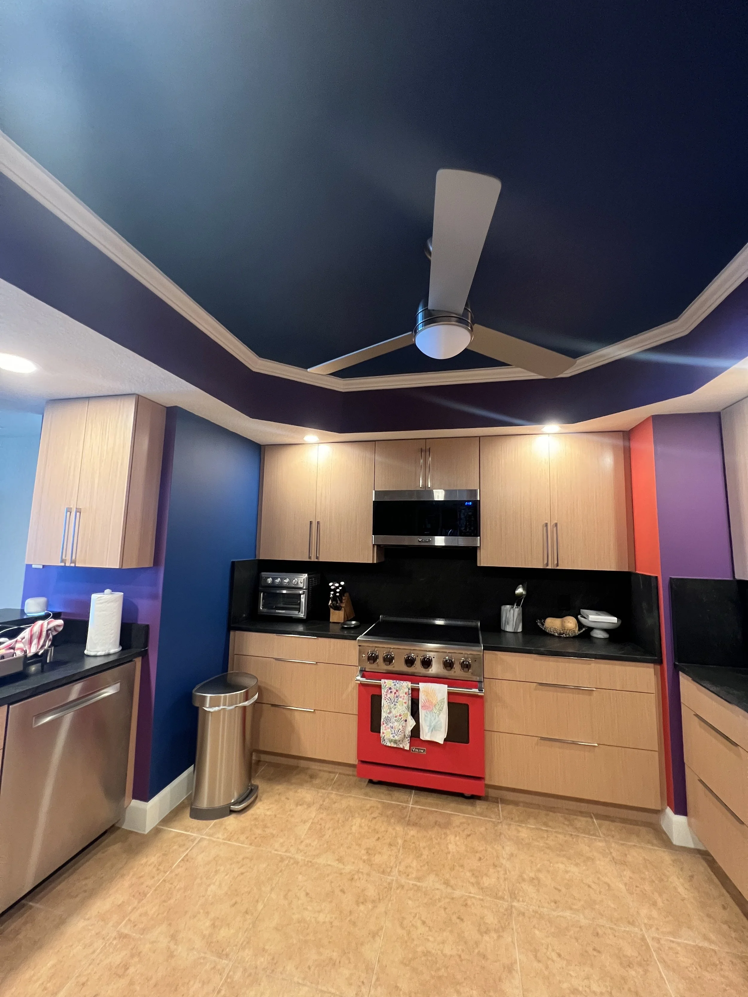 A colorful kitchen with a blue ceiling, beige cabinets, red oven, stainless steel trash can, and various small appliances and kitchen items on the countertops.