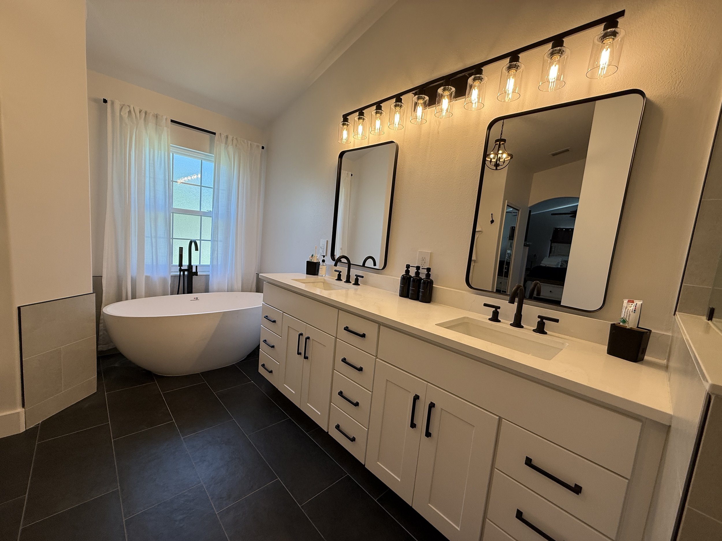 A new custom modern bathroom remodel, featuring a white double vanity with black handles, two mirrors, black faucets, a freestanding bathtub, and a wall-mounted light fixture with custom tiling on the floors and walls. 