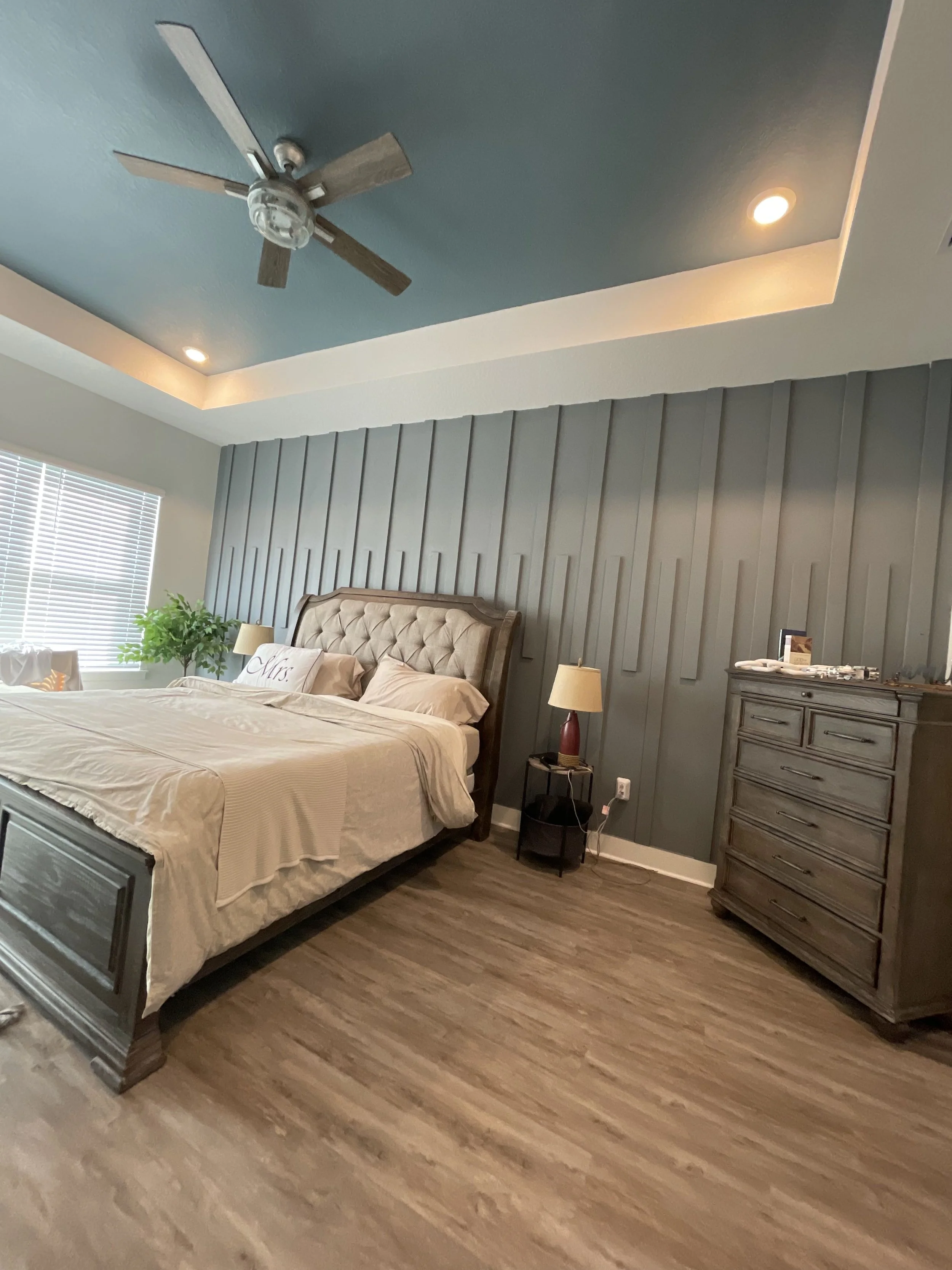 A bedroom with a bed, beige bedding and pillows, a gray wooden dresser, a nightstand with a lamp, a window with blinds, a ceiling fan, and wood-look flooring.