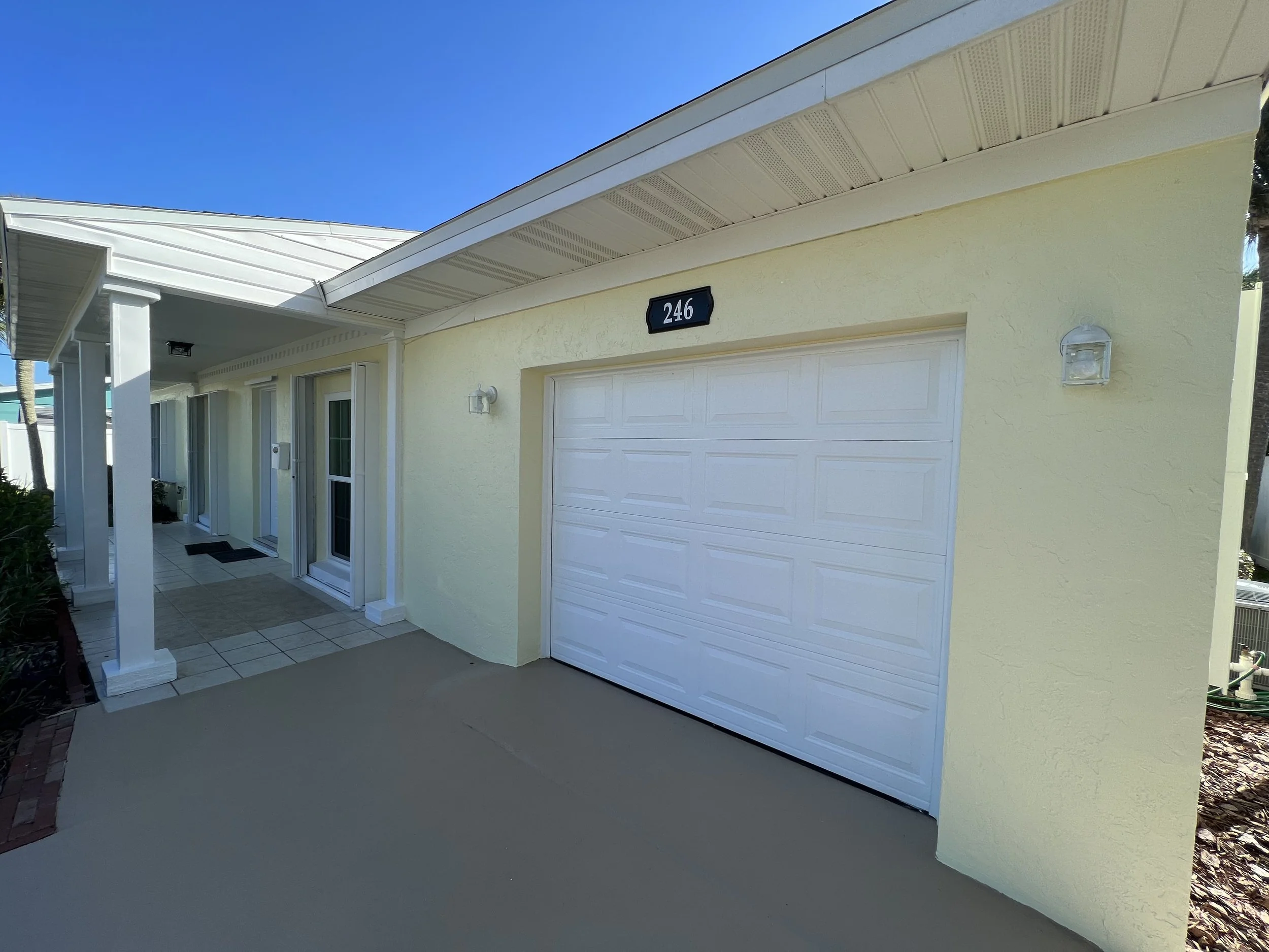 Front view of a yellow house with a white garage door, house number 246, and a porch with white posts, located under a clear blue sky.