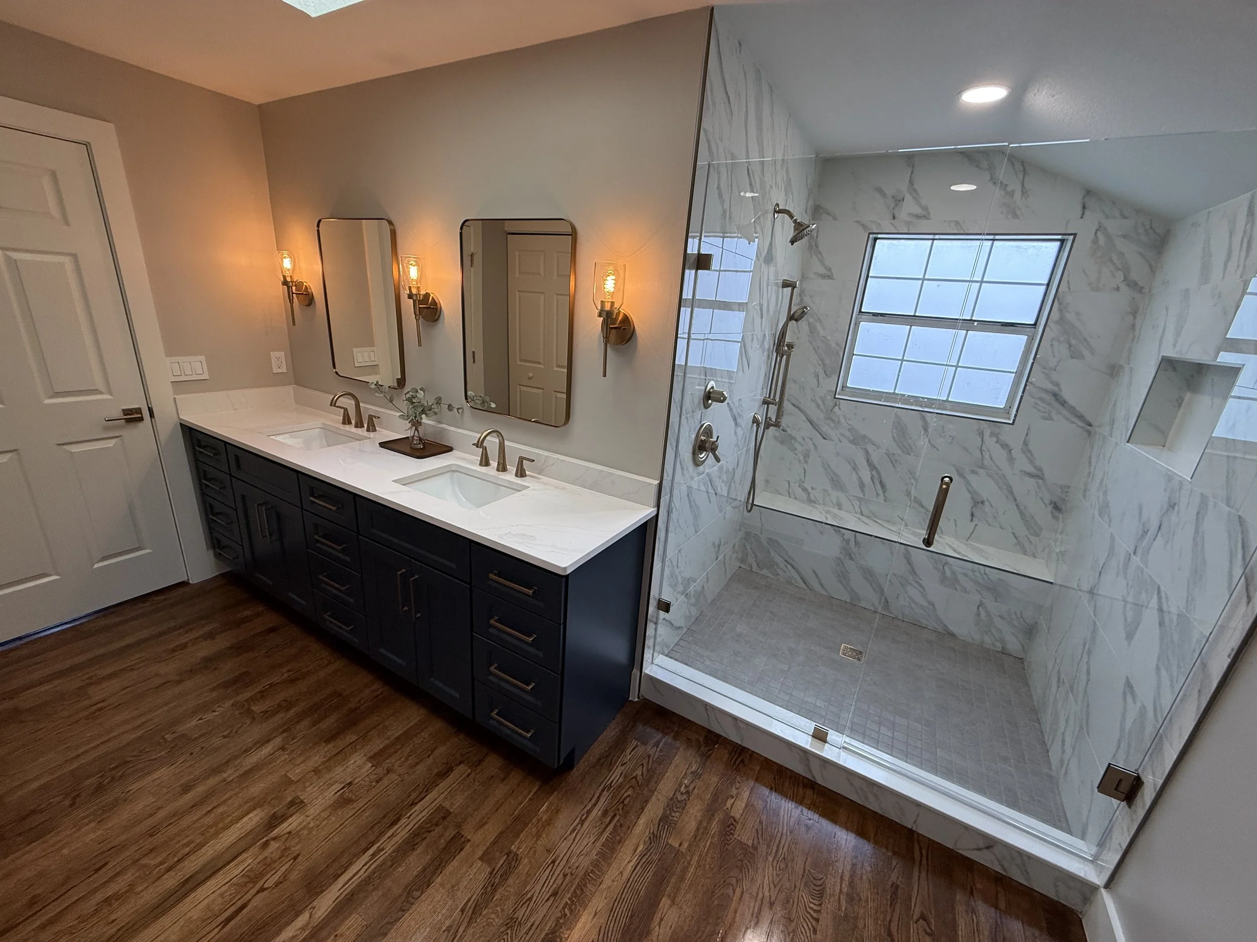 Modern bathroom with dual sinks, mirrors, wall sconces, gray cabinets, wood flooring, and a glass-enclosed shower with marble walls and windows.