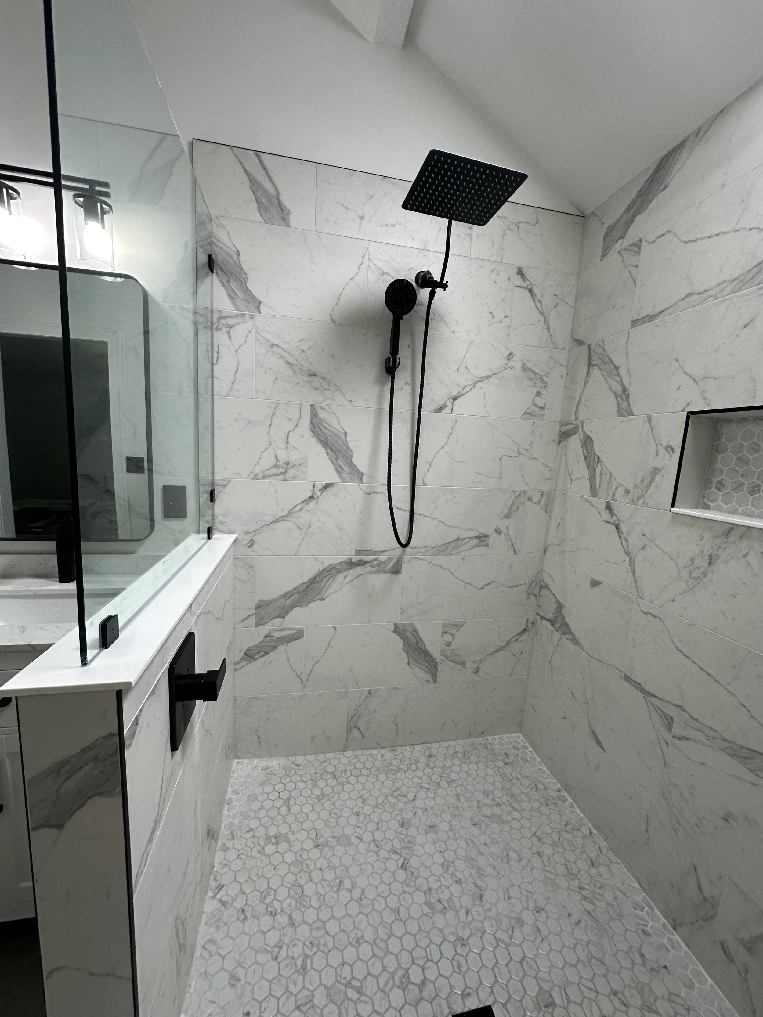 A modern shower area with a black overhead rain shower and handheld shower head mounted on a marble wall. The shower floor has small honeycomb tiles, and there's a built-in niche with hexagonal tiles. A partial glass shower partition is visible on the left.