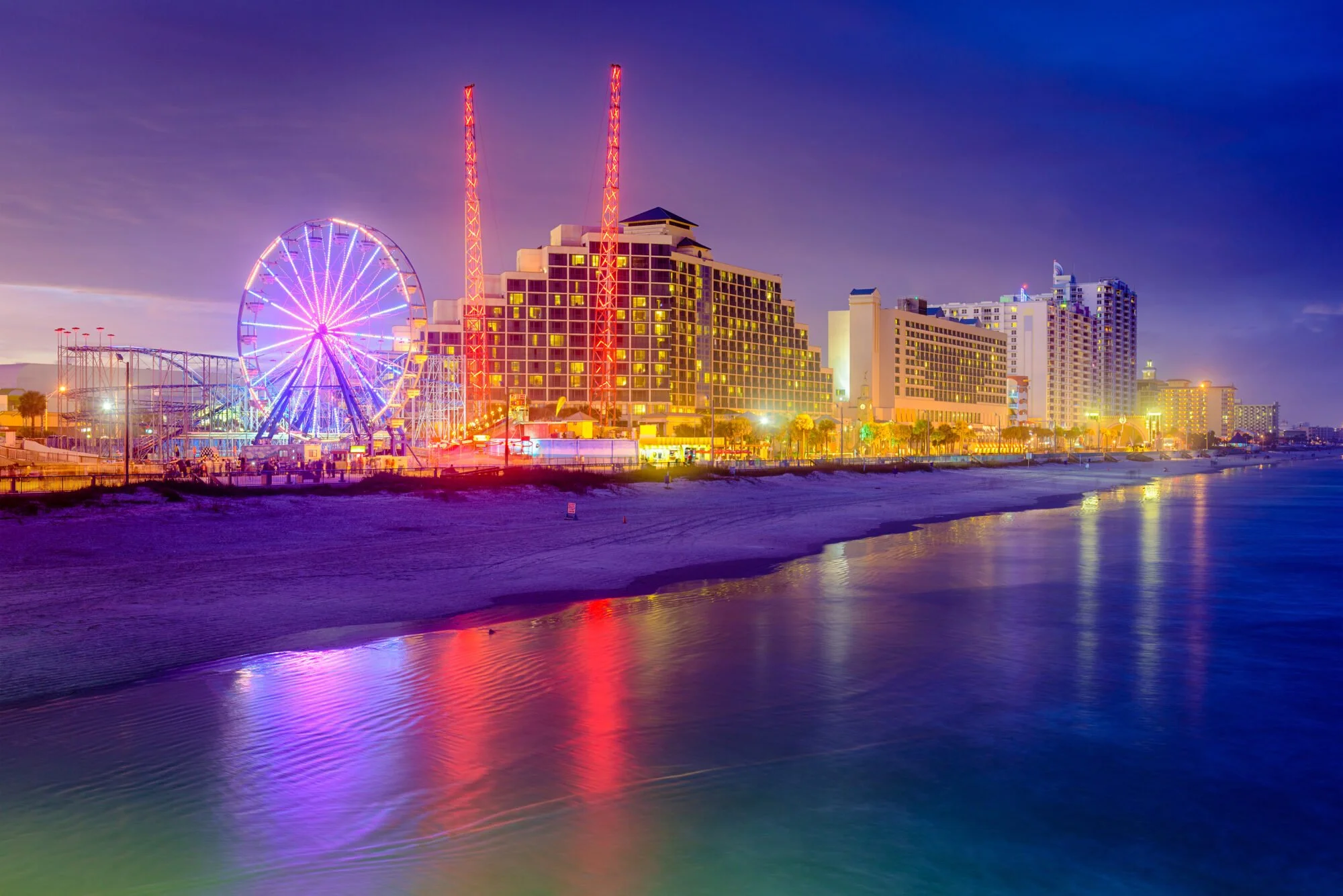 Nighttime view of a lively beachside city with brightly lit buildings, a Ferris wheel, and roller coaster, reflecting colorful lights on the water.