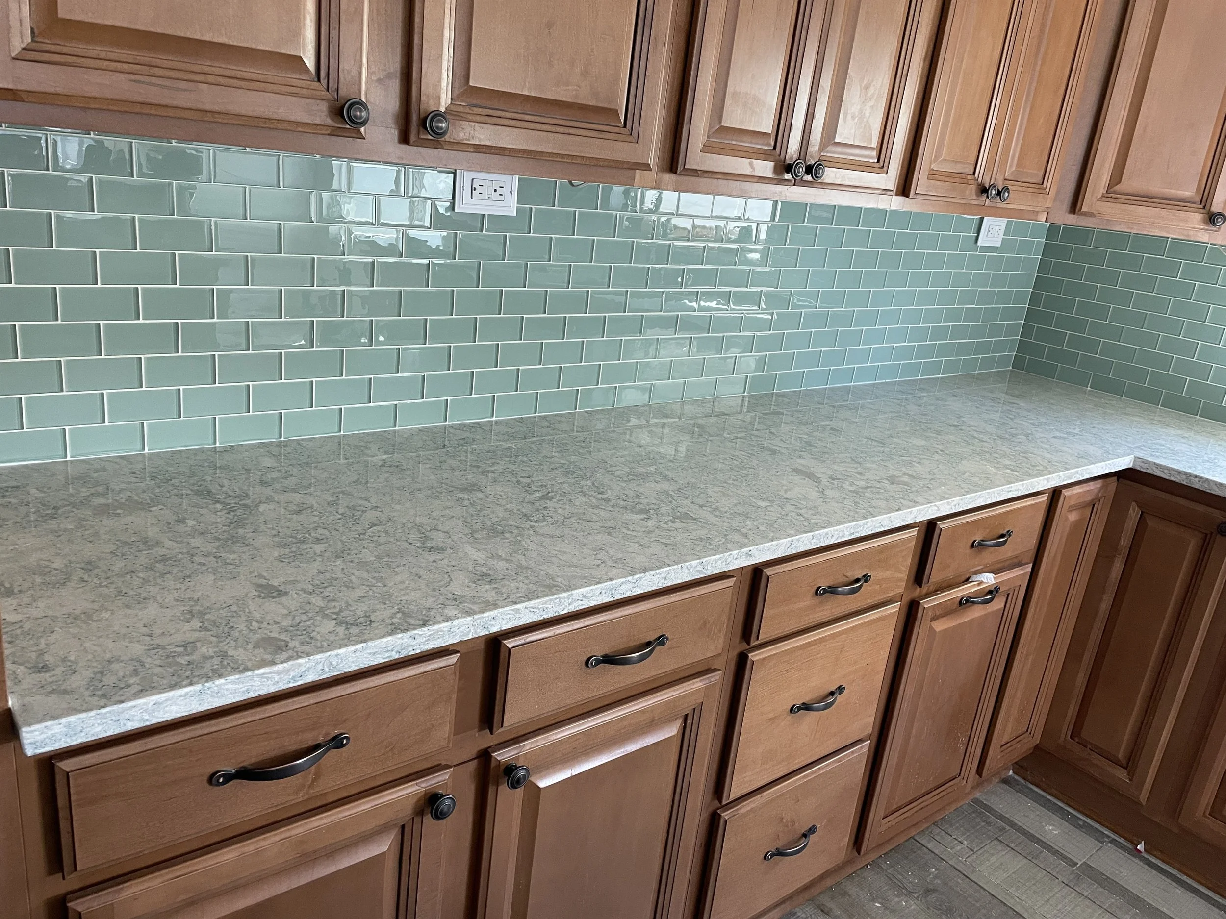 A kitchen countertop with a light-colored stone surface, wooden cabinets with black handles, mint green subway tile backsplash, and two white electrical outlets.