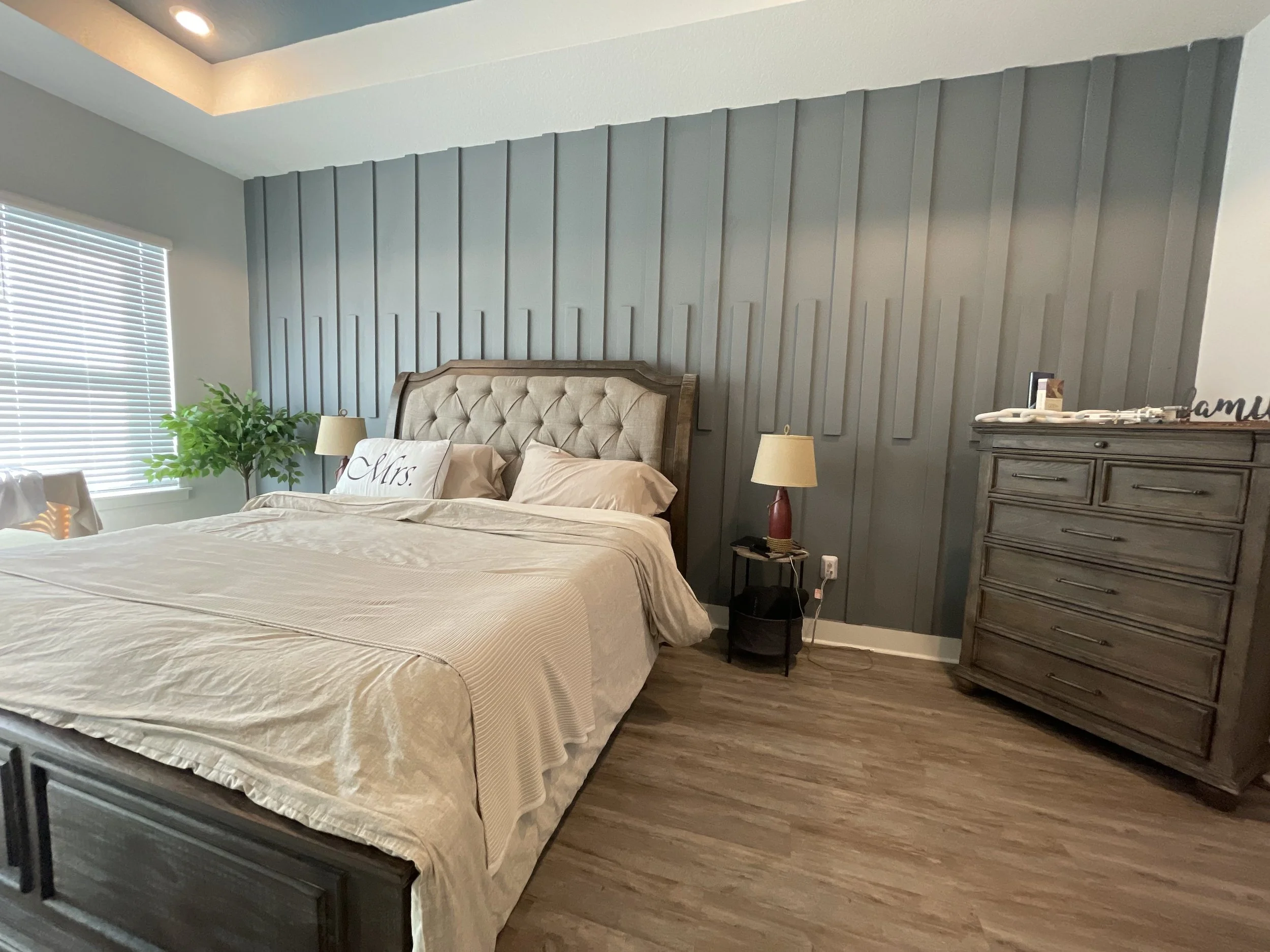 A bedroom with a large bed, beige bedding, and decorative pillows, including one with the word 'Mrs.' on it. There is a wooden headboard, two bedside lamps, a plant, and a gray dresser. The wall behind the bed features gray vertical paneling. A window with white blinds is on the left side, and a small table with items on top and a power outlet with cords is on the right side of the bed.