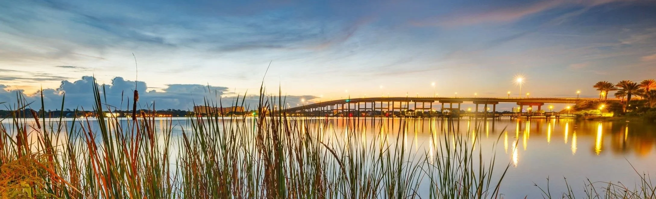 Sunset over a calm body of water with a bridge and streetlights, tall grass in the foreground, and cloudy sky.
