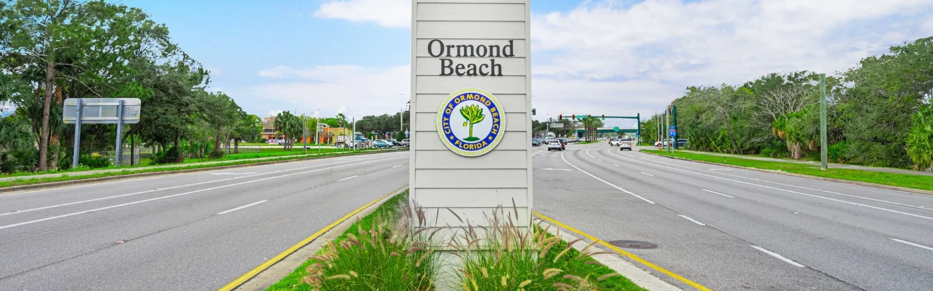 Sign indicating Ormond Beach with the city seal of City of Ormond Beach, Florida, on a road with cars and trees in the background.