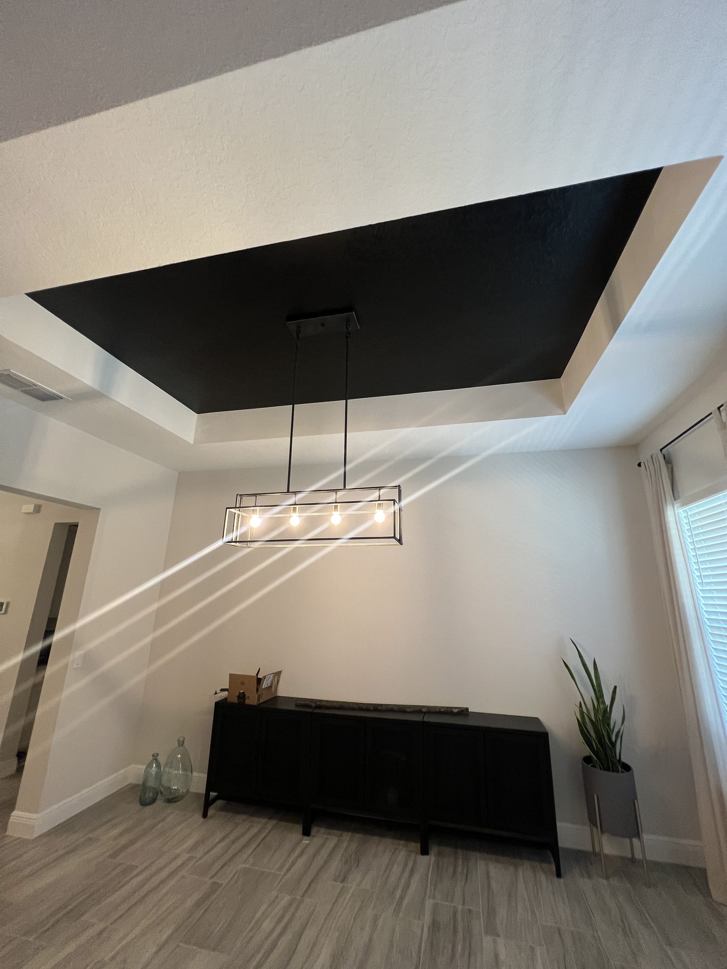 A modern living room ceiling with a black recessed lighting panel and a rectangular chandelier with exposed bulbs. There is a black sideboard below the ceiling, with glass bottles and a potted plant nearby. Part of a window with white blinds and a curtain can be seen on the right.