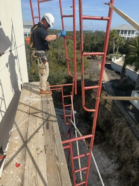 A construction worker wearing a helmet, gloves, and safety harness standing on a wooden platform, installing or inspecting red metal scaffolding outside a building on a sunny day.
