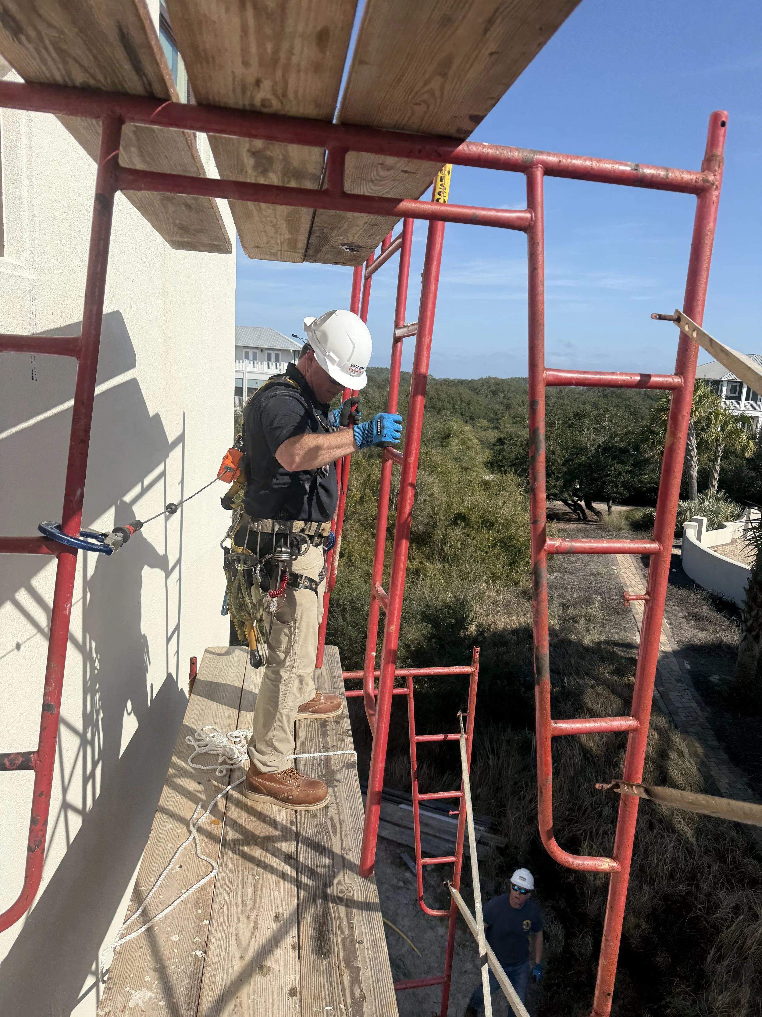 Construction worker wearing a white helmet and black shirt using tools on a scaffold outside a building, with another worker visible below.