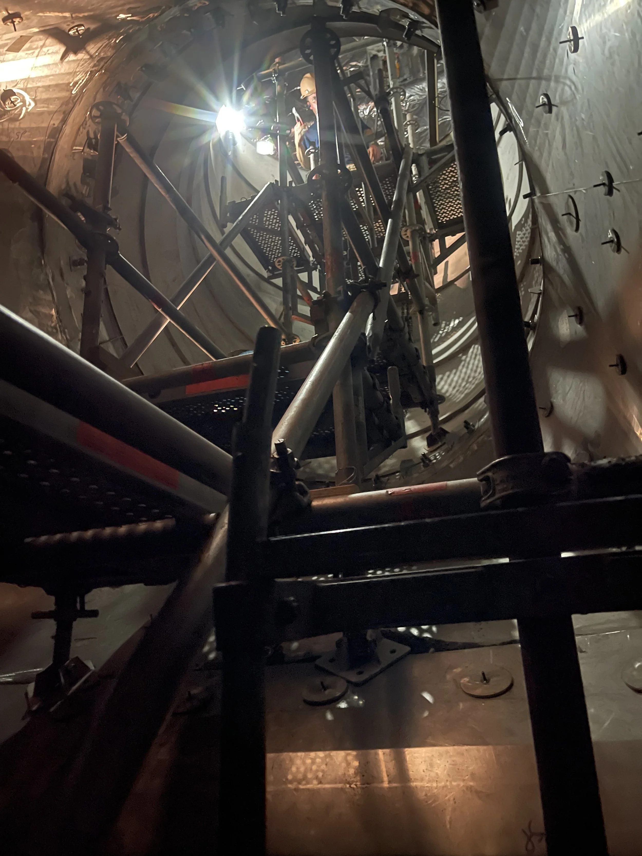 Inside a tunnel boring machine, showing the internal machinery, metal framework, and a worker seated at the control panel, with bright light shining through.