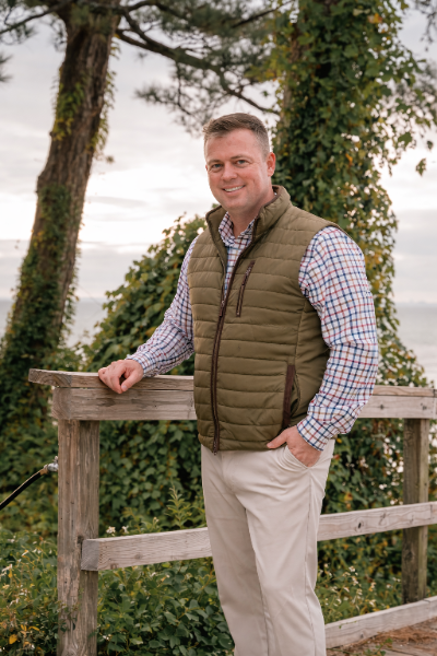 A smiling man standing outdoors on a wooden railing, with trees and a body of water in the background.