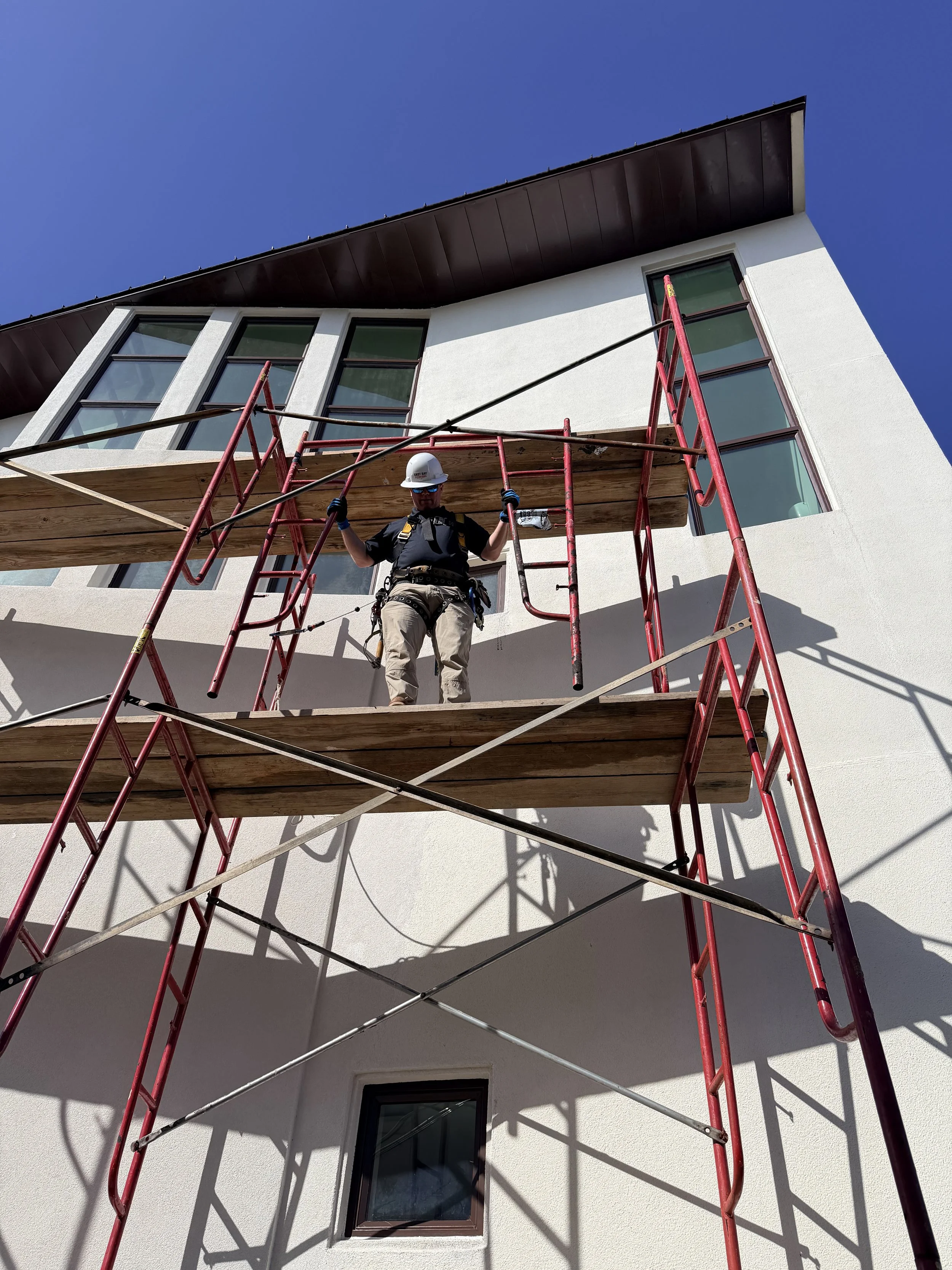 Construction worker standing on scaffolding outside a white building, wearing a safety helmet and harness, under a clear blue sky.