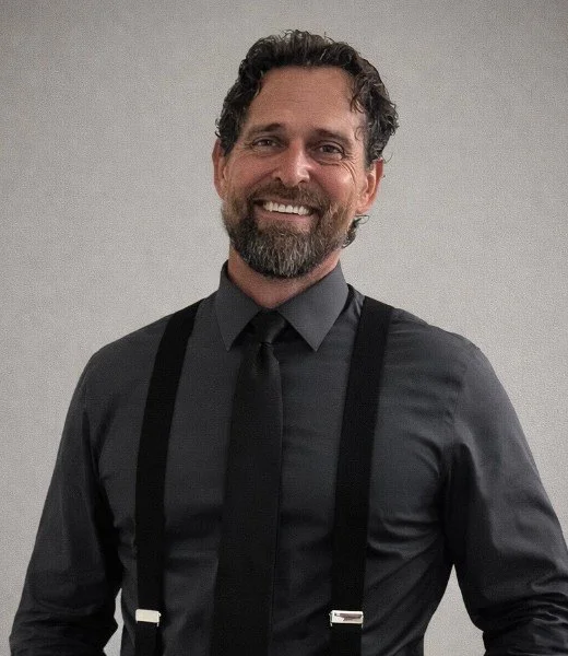 A smiling man with dark curly hair and a beard, wearing a black shirt, black suspenders, and a black tie, standing against a plain gray background.