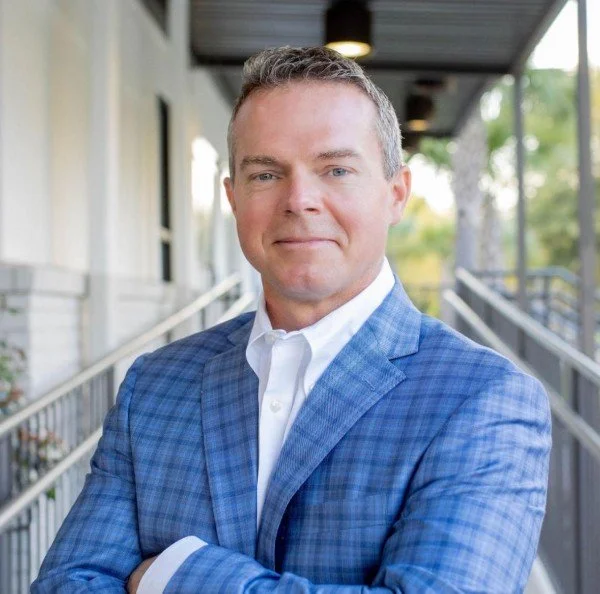 A man in a blue plaid suit jacket and white shirt standing outside on a balcony or walkway, with a building and trees in the background.