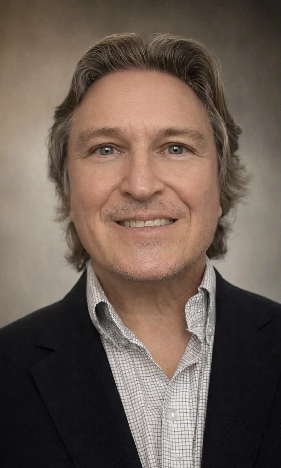 A professional headshot of a smiling middle-aged man with blue eyes, wavy light brown hair, wearing a dark suit jacket and a light patterned button-up shirt against a neutral background.