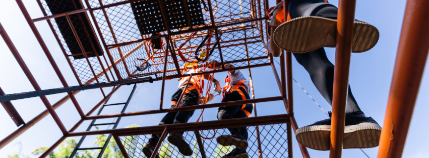 Two people in safety gear climbing a metal jungle gym outdoors under a clear blue sky.