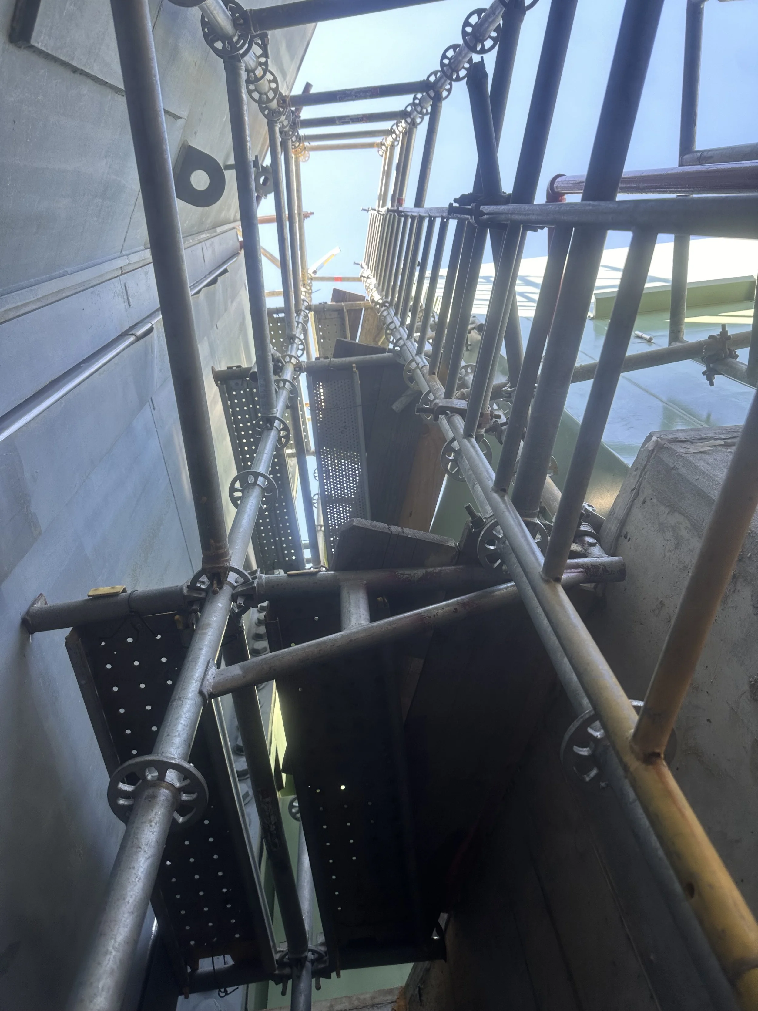 Scaffolding on a building under construction, with metal pipes, wheels, and a platform, viewed from below against a blue sky.