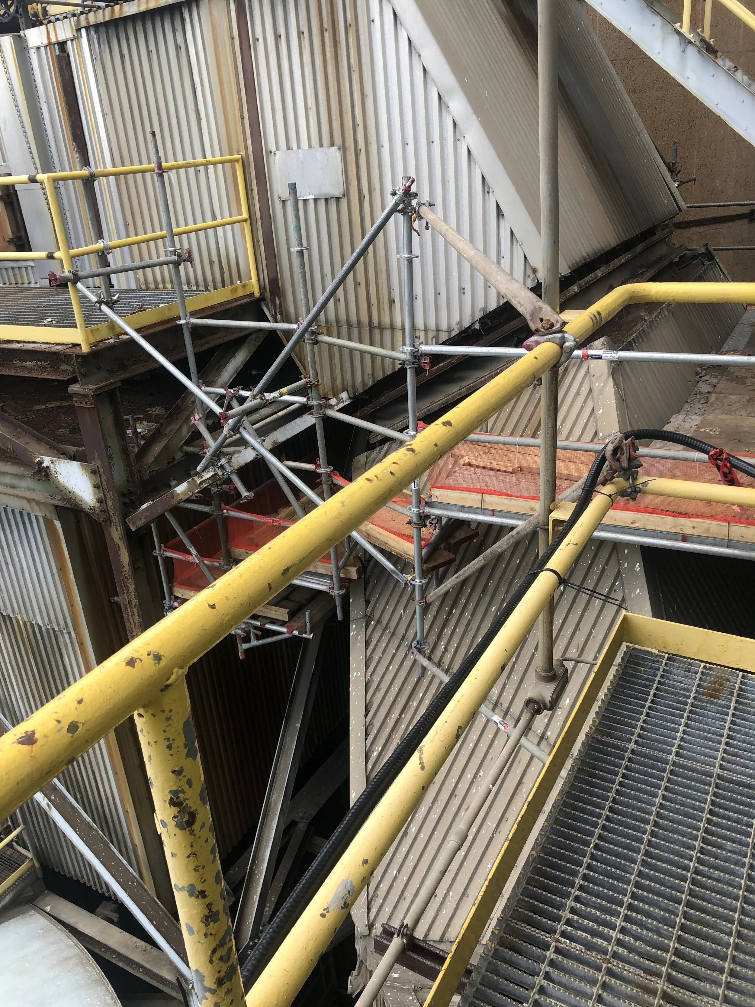 A construction site with metal scaffolding, yellow safety railings, and corrugated metal walls and stairs.
