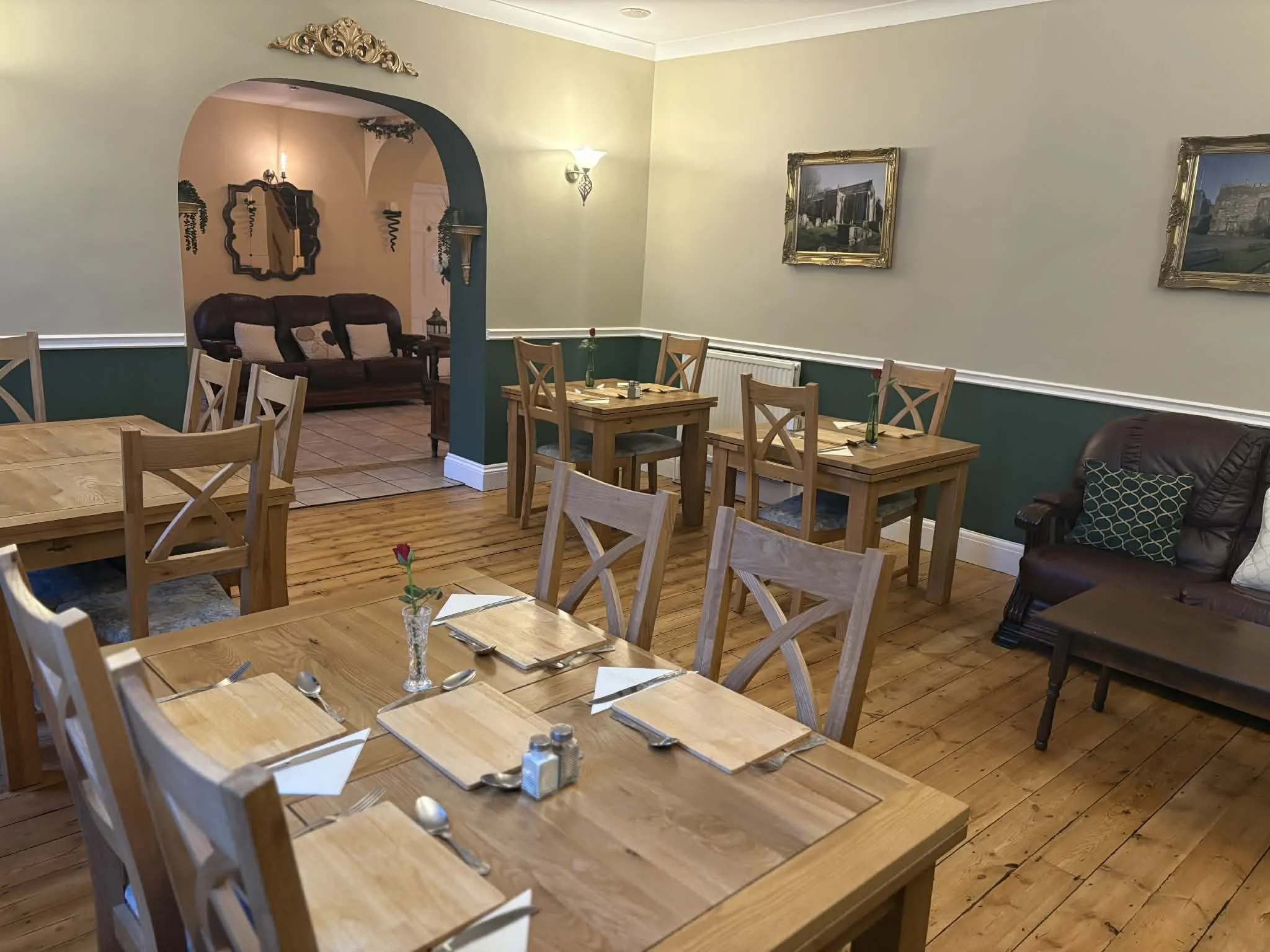 Cozy dining area with wooden tables and chairs, set with placemats, napkins, and utensils, with a small flower vase on one table, and art framed pictures on the walls.