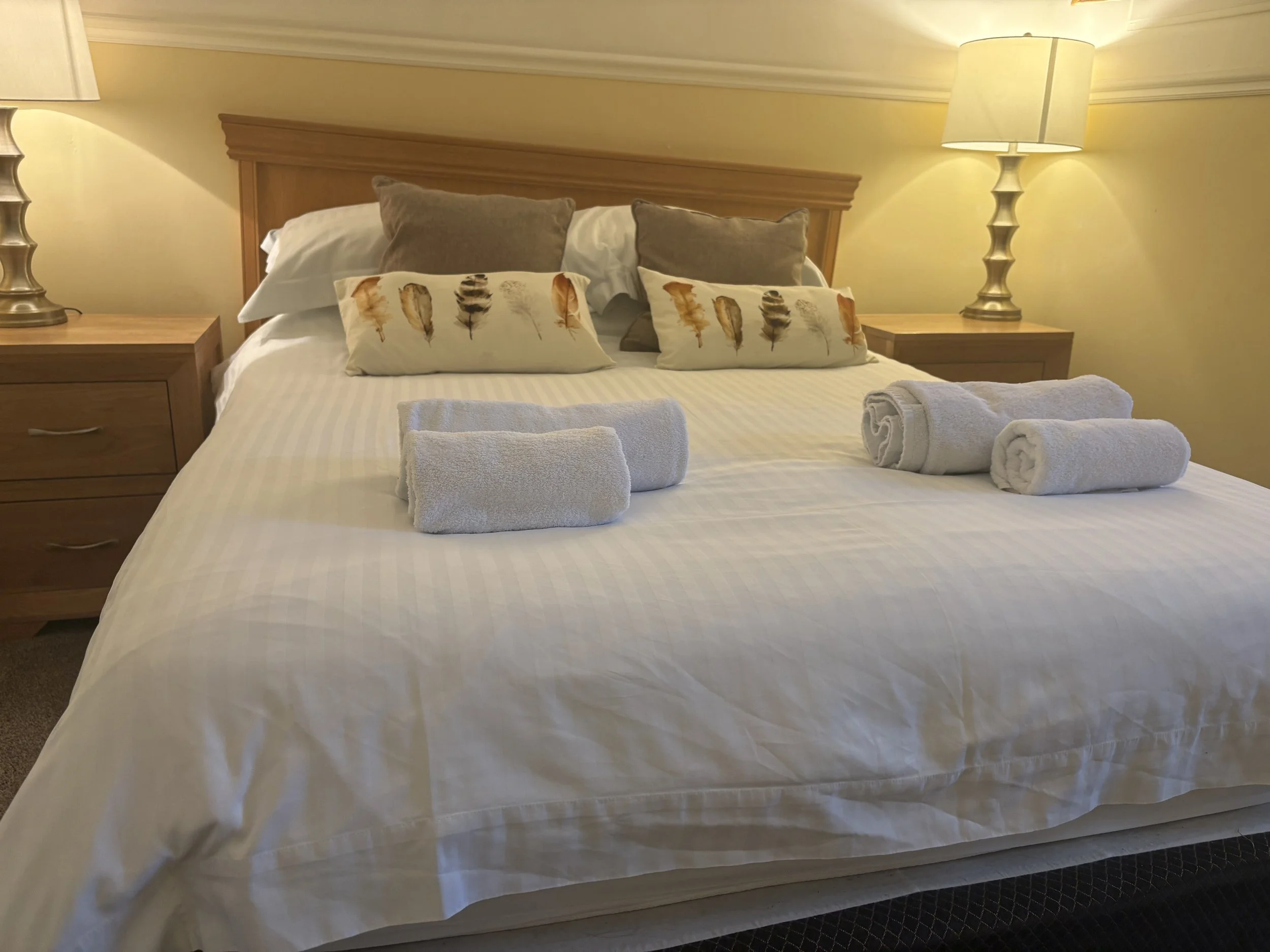 A neatly made bed with white bedding, two gray pillows, three white decorative pillows with feather prints, and three rolled white towels on top. There are wooden nightstands with lamps on each side of the bed, and a wooden headboard.