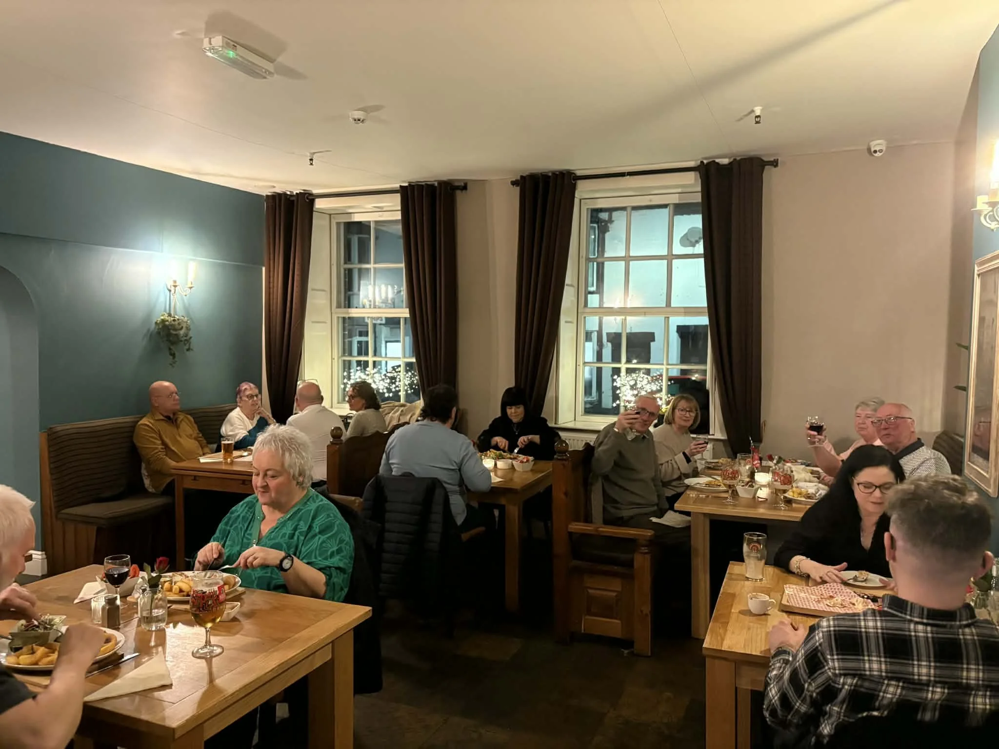 People dining in a warmly lit restaurant with blue and beige walls, large windows with brown curtains, and holiday decorations visible outside through the windows.