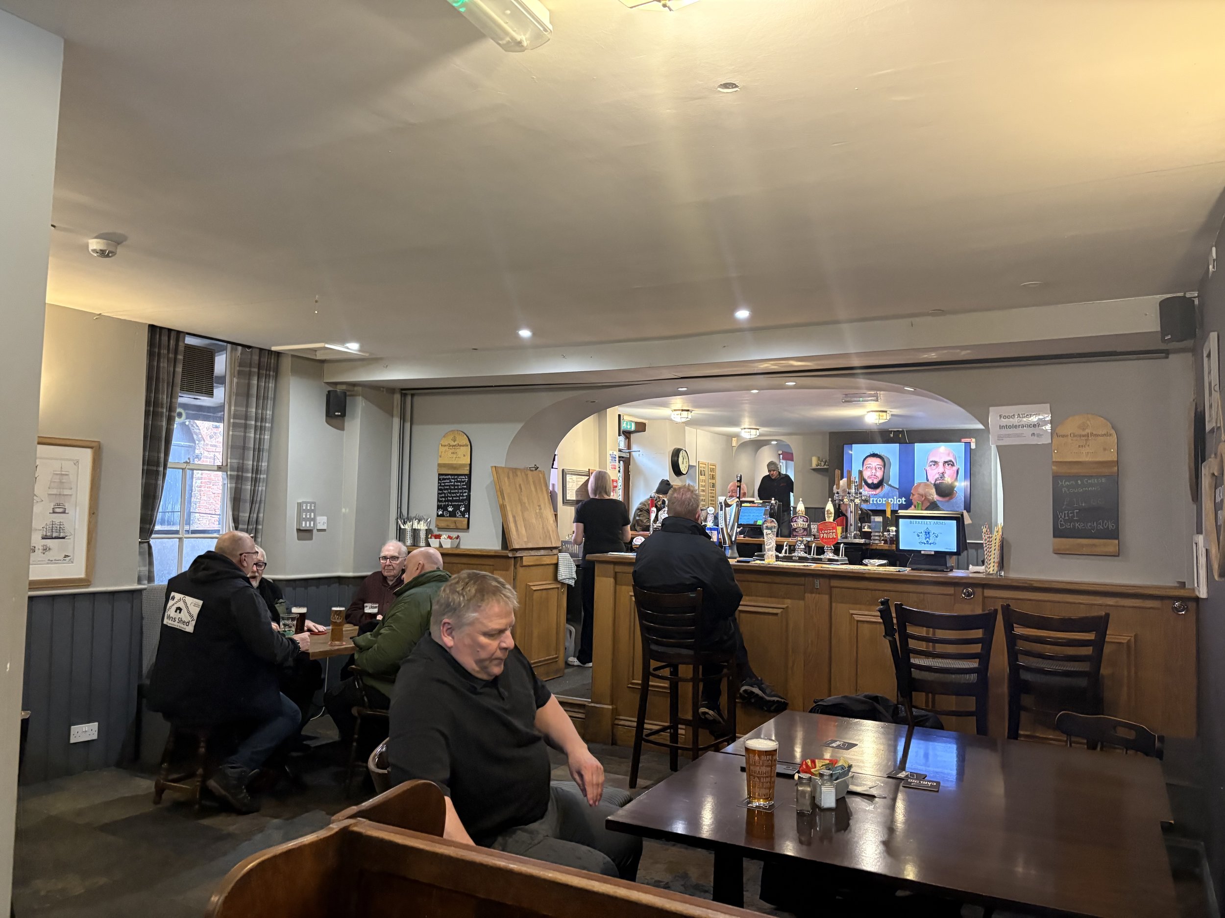 A cozy pub with a bar area in the back, several patrons sitting at tables, watching sports on TV, and enjoying drinks. The decor features wooden furniture, checkered curtains, and framed pictures on the walls.