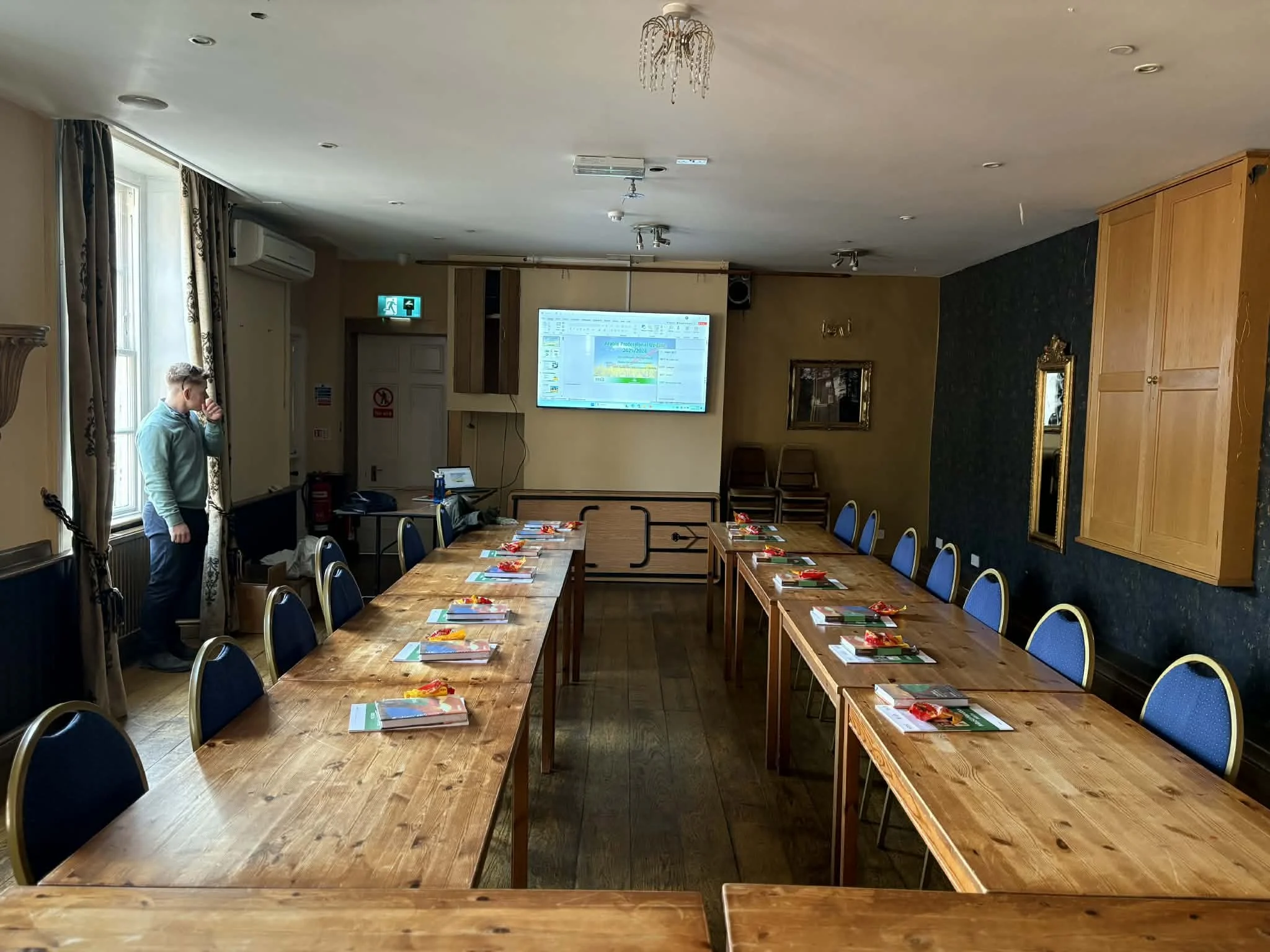 Empty conference room with two long wooden tables set with notebooks and red pens, blue chairs, a man standing near the window, a large screen at the front, and a chandelier hanging from the ceiling.