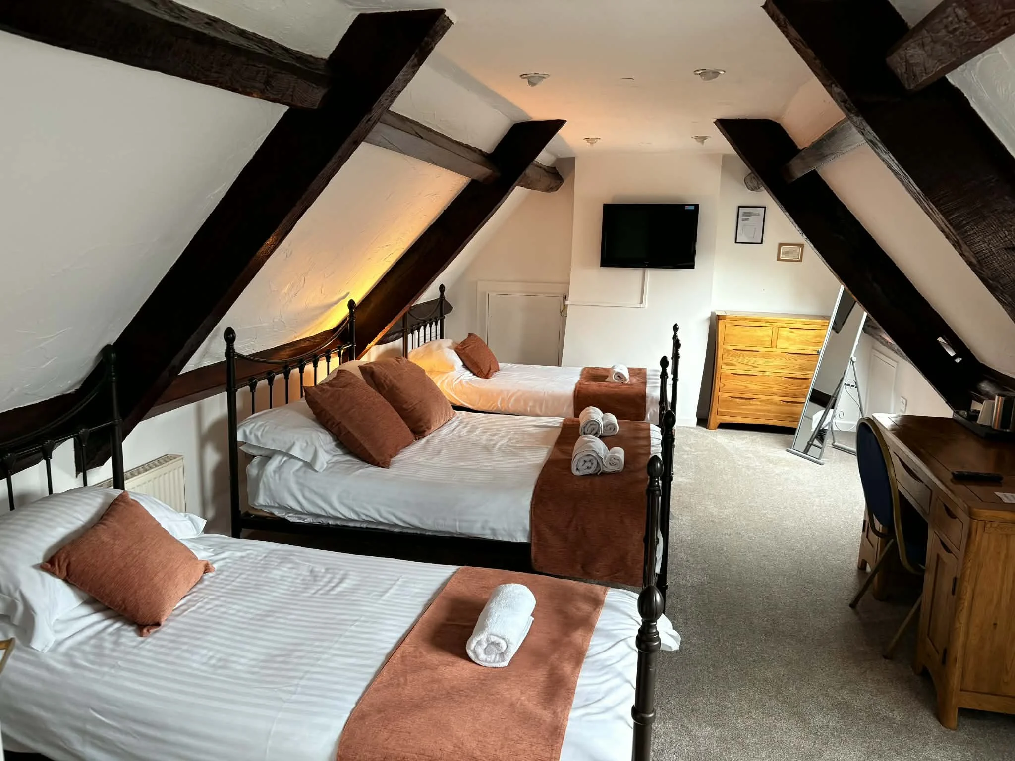 A cozy hotel room with three metal framed beds, each with white sheets, brown pillows, and rolled towels. The room has a sloped ceiling with dark wooden beams. There is a small wooden dresser, a flat-screen TV on the wall, a mirror, and a wooden desk
