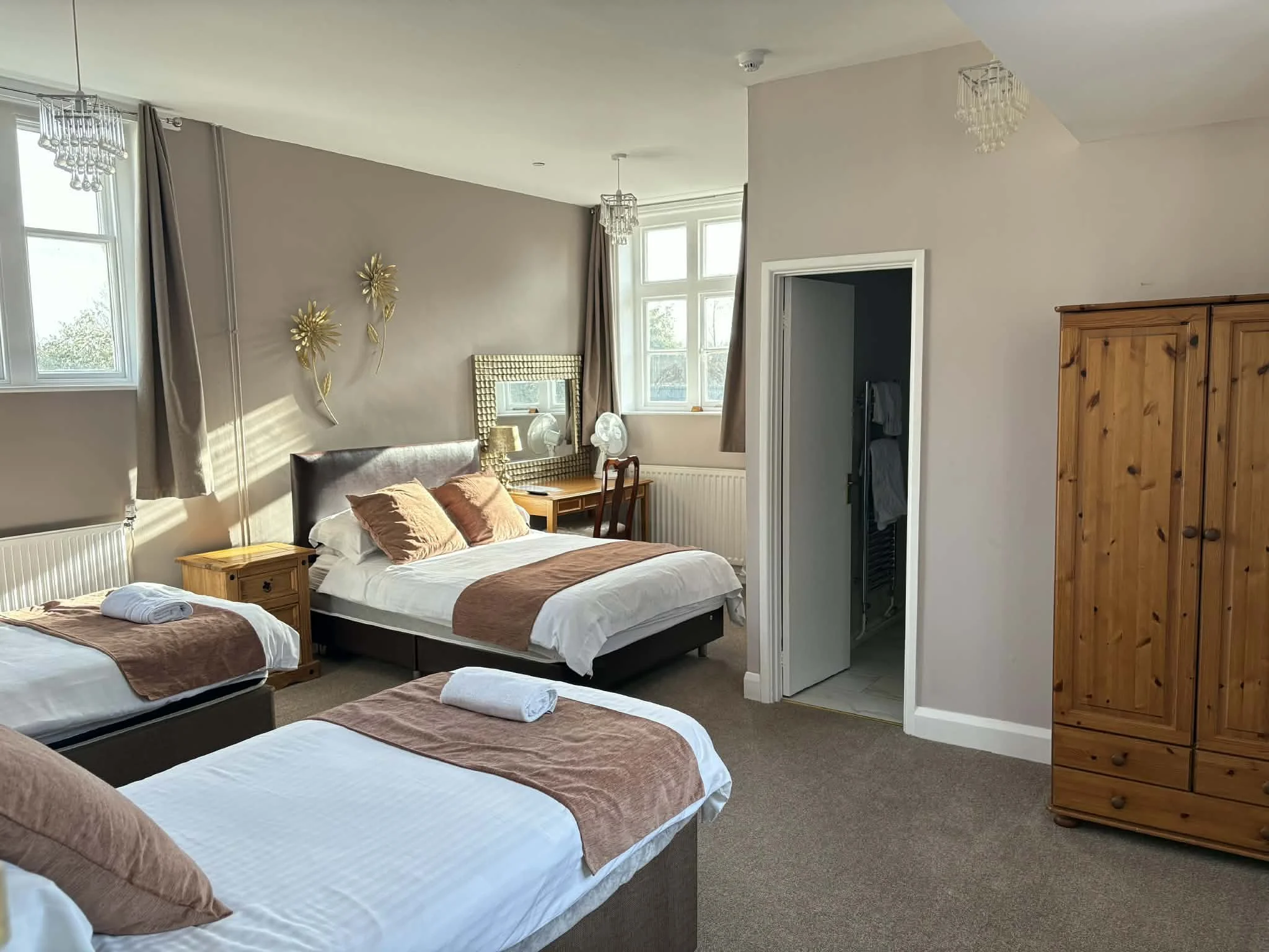 Photo of a bedroom with two single beds and one large bed, all made with white sheets and brown bed runners, towels placed at the foot of each bed, wooden bedside tables, a wooden desk with a chair, mirror, lamps, golden wall decorations, large windo