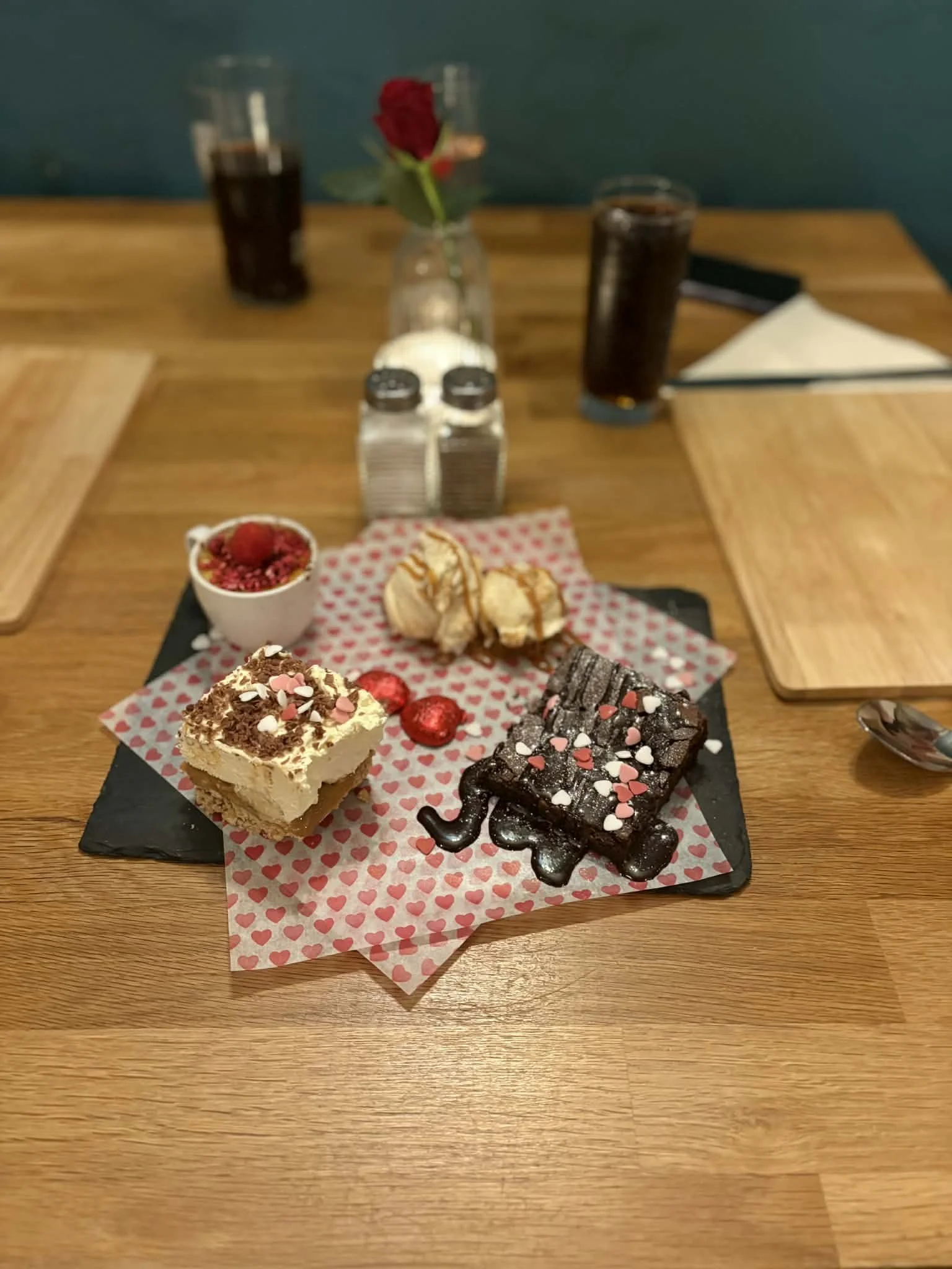 Valentine's Day dessert platter with various sweets, including a chocolate brownie decorated with heart-shaped sprinkles, a cake slice with a layer of chocolate, a small cup of raspberries and red berries, and a few pieces of croissants, on a black r