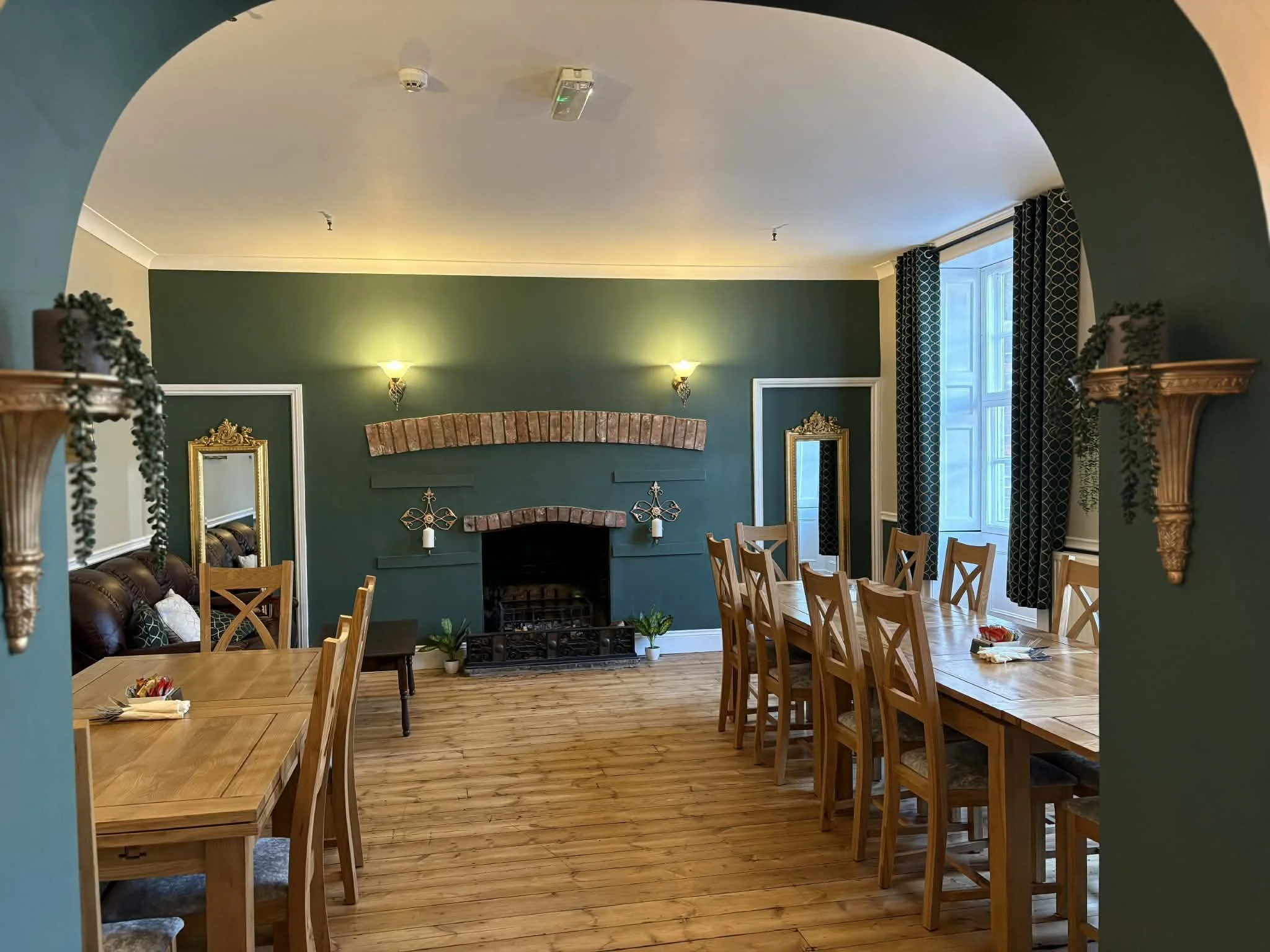 Elegant dining room with green walls, a brick fireplace, and wooden tables and chairs. Decor includes gold-framed mirrors and greenery, with a large window and dark patterned curtains.