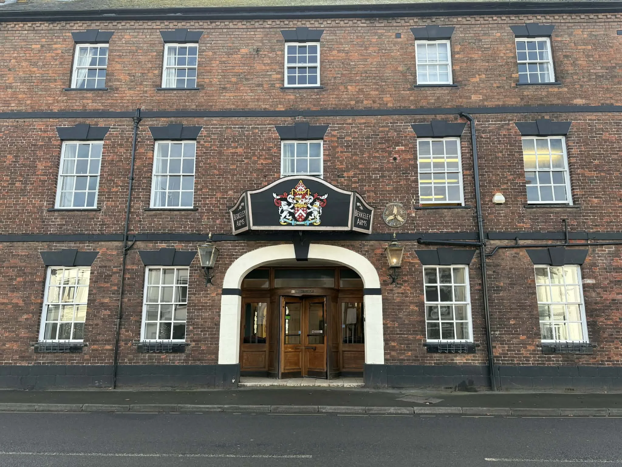 Exterior view of the Berkeley Arms pub, a brick building with multiple windows and a central wooden entrance door, above which is a crest and sign.