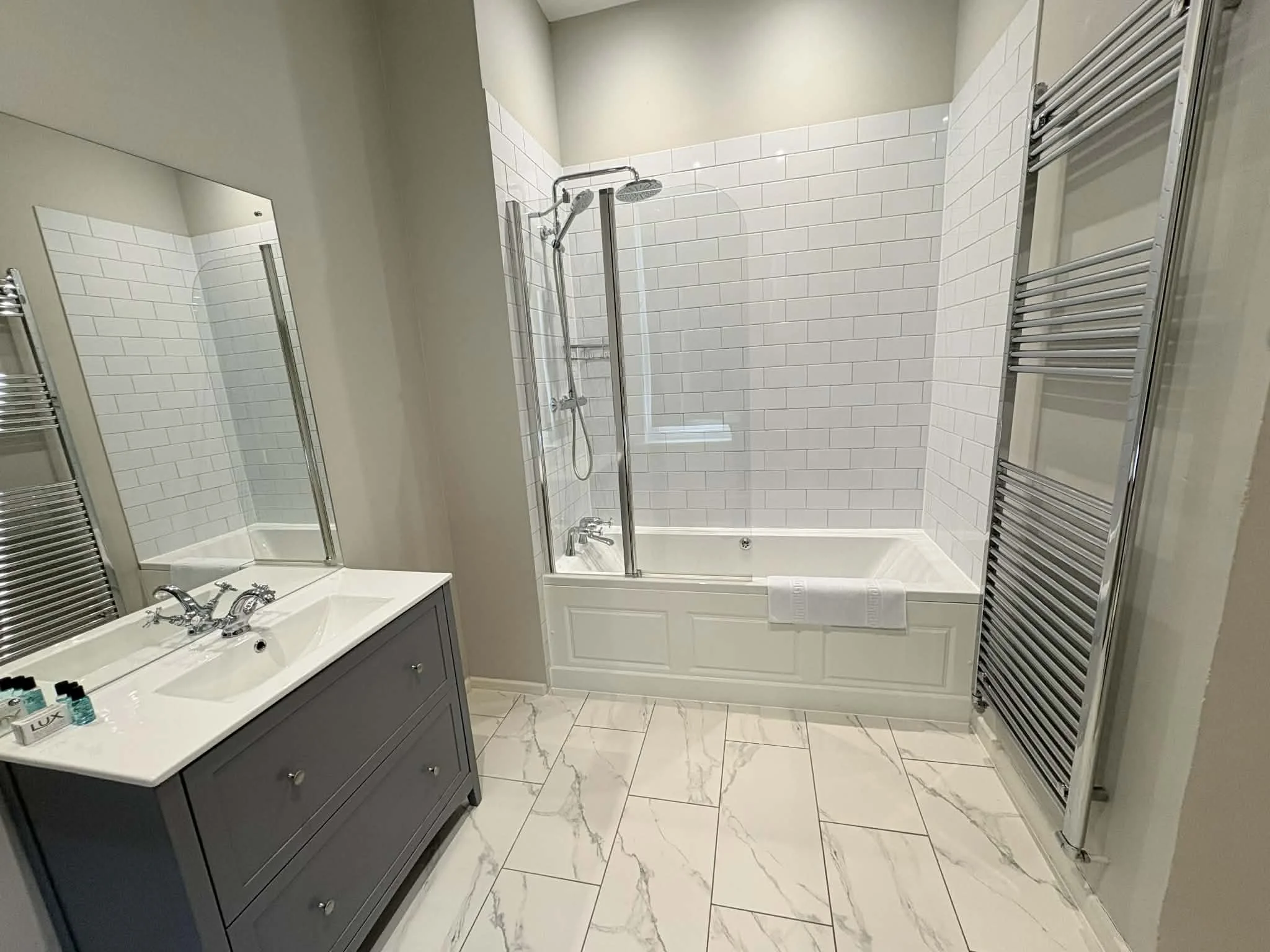 Bathroom with a double sink vanity, large mirror, bathtub with glass partition, and wall-mounted towel radiators. White subway tiles on the wall and marble-patterned floor tiles.