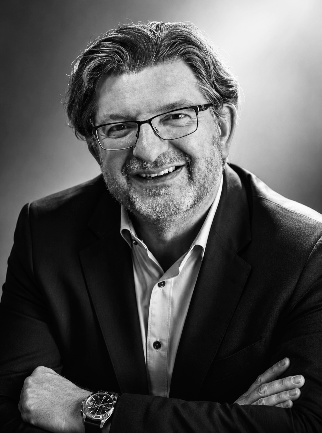 Black-and-white portrait of a smiling middle-aged man with glasses, a beard, and shoulder-length hair, wearing a suit jacket, shirt, and a watch on his left wrist, with arms crossed.