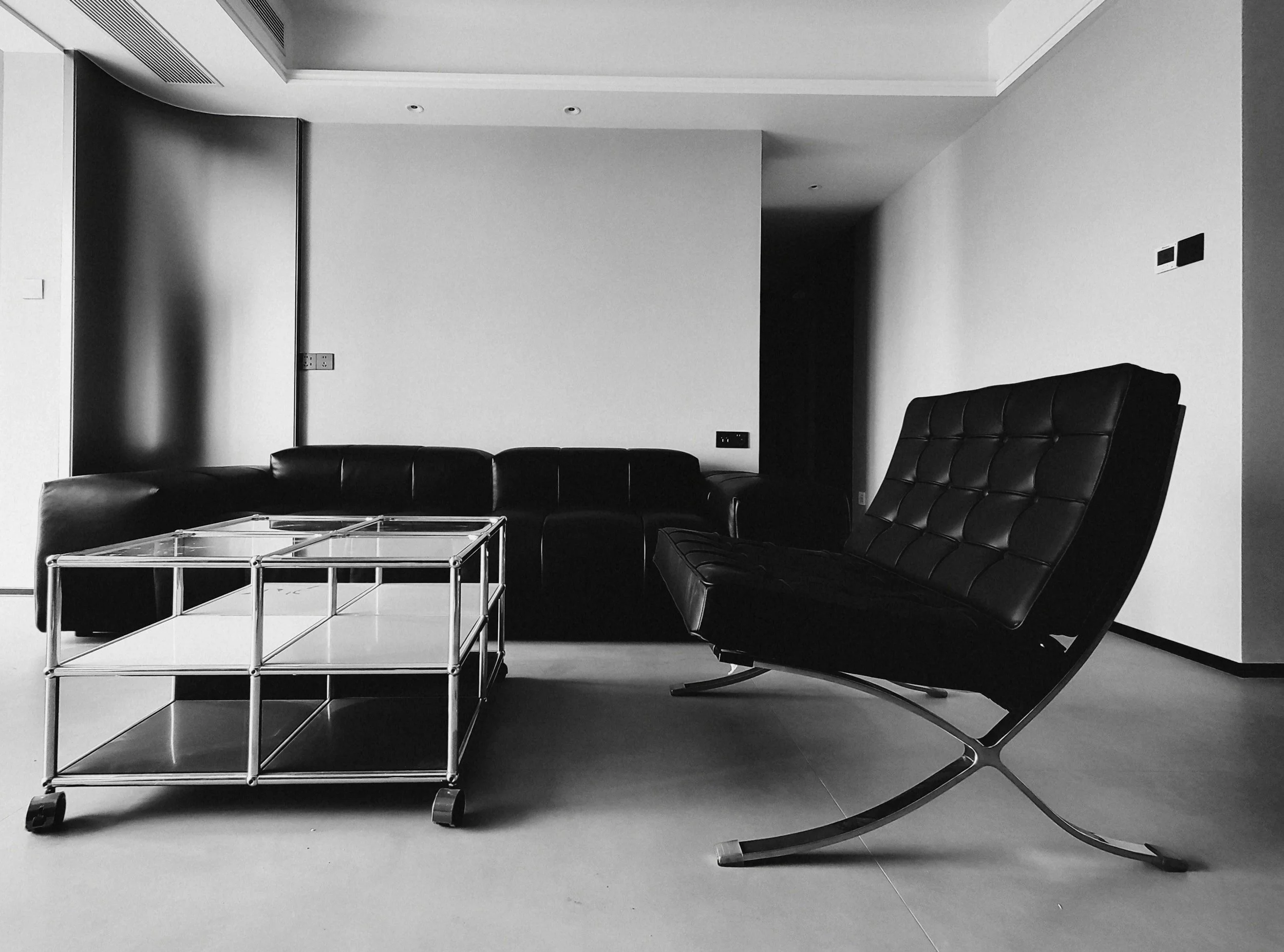 Modern living room with a black leather sofa and a glass coffee table on a gray floor, with white walls and a ceiling with recessed lighting.