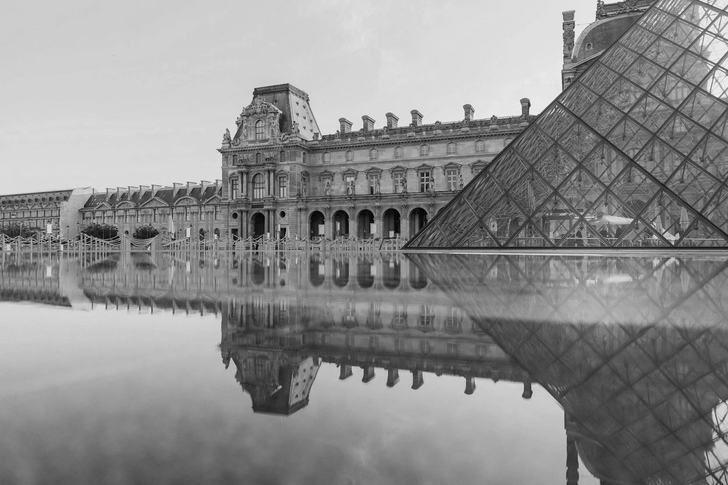 Black and white photo of the Louvre Museum with its glass pyramid entrance, reflected in a puddle on the ground.