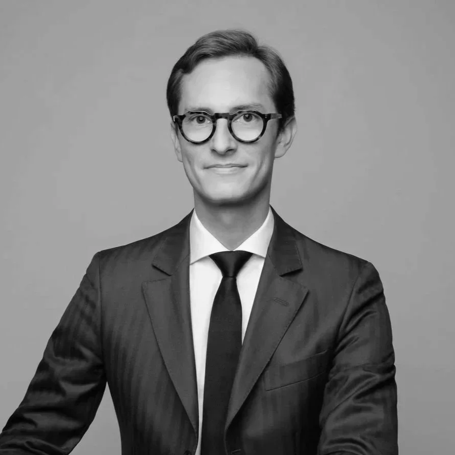 Black and white professional portrait of a man wearing a suit, tie, and round glasses, smiling slightly against a plain background.