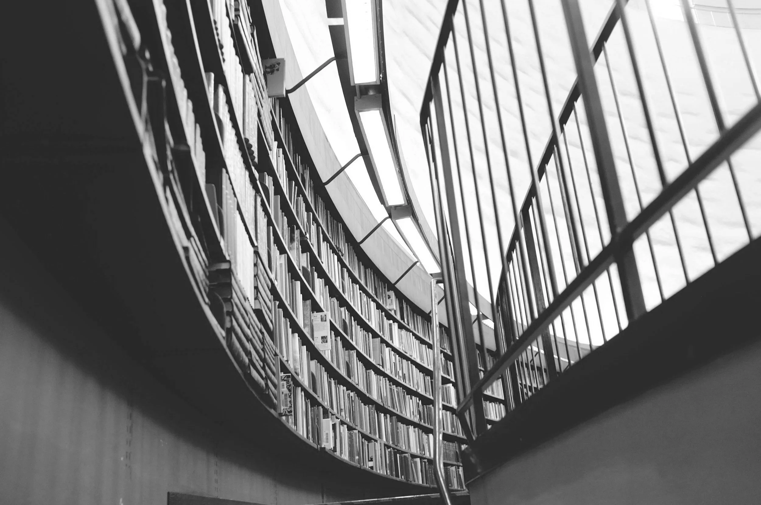 Black and white photo of a modern interior with tall bookshelves filled with books seen from below, with railing on the side.