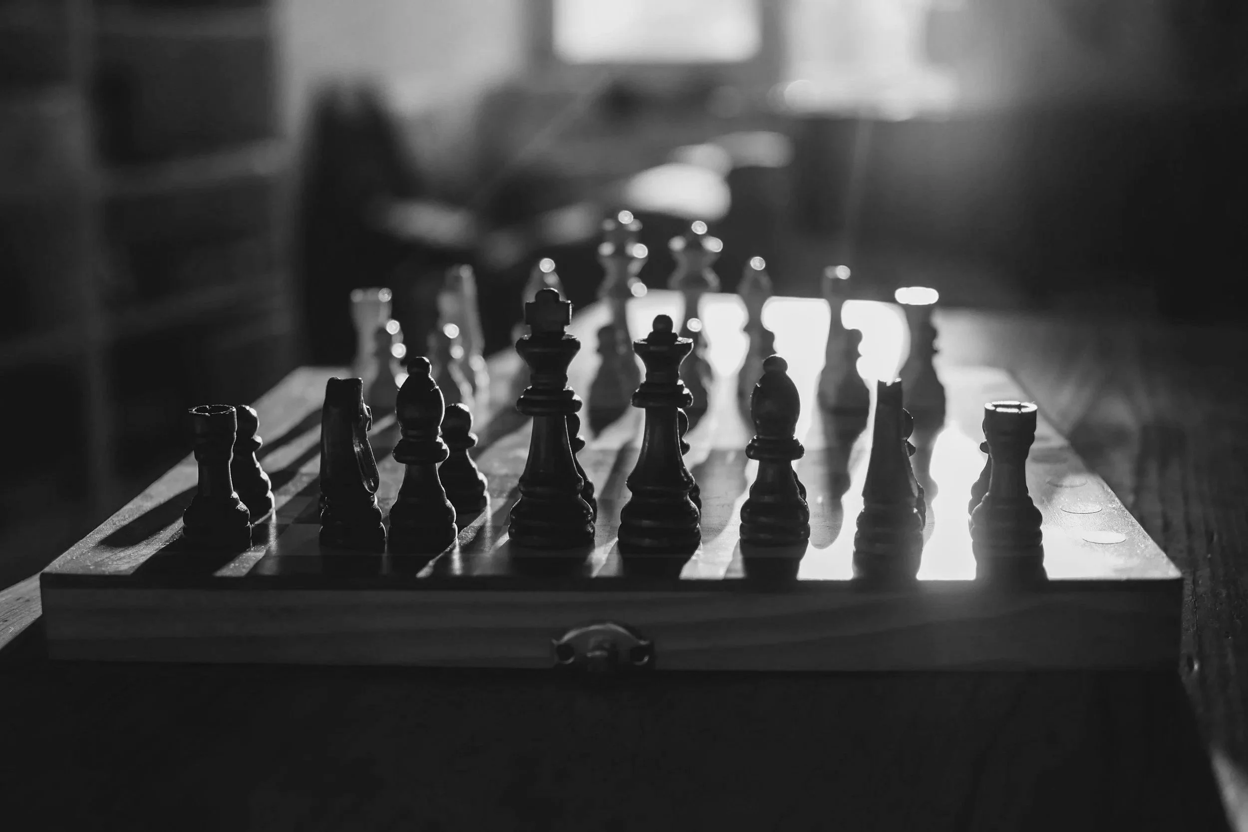 Black and white close-up of a chessboard with chess pieces arranged for a game, sunlight streaming in through a window in the background.