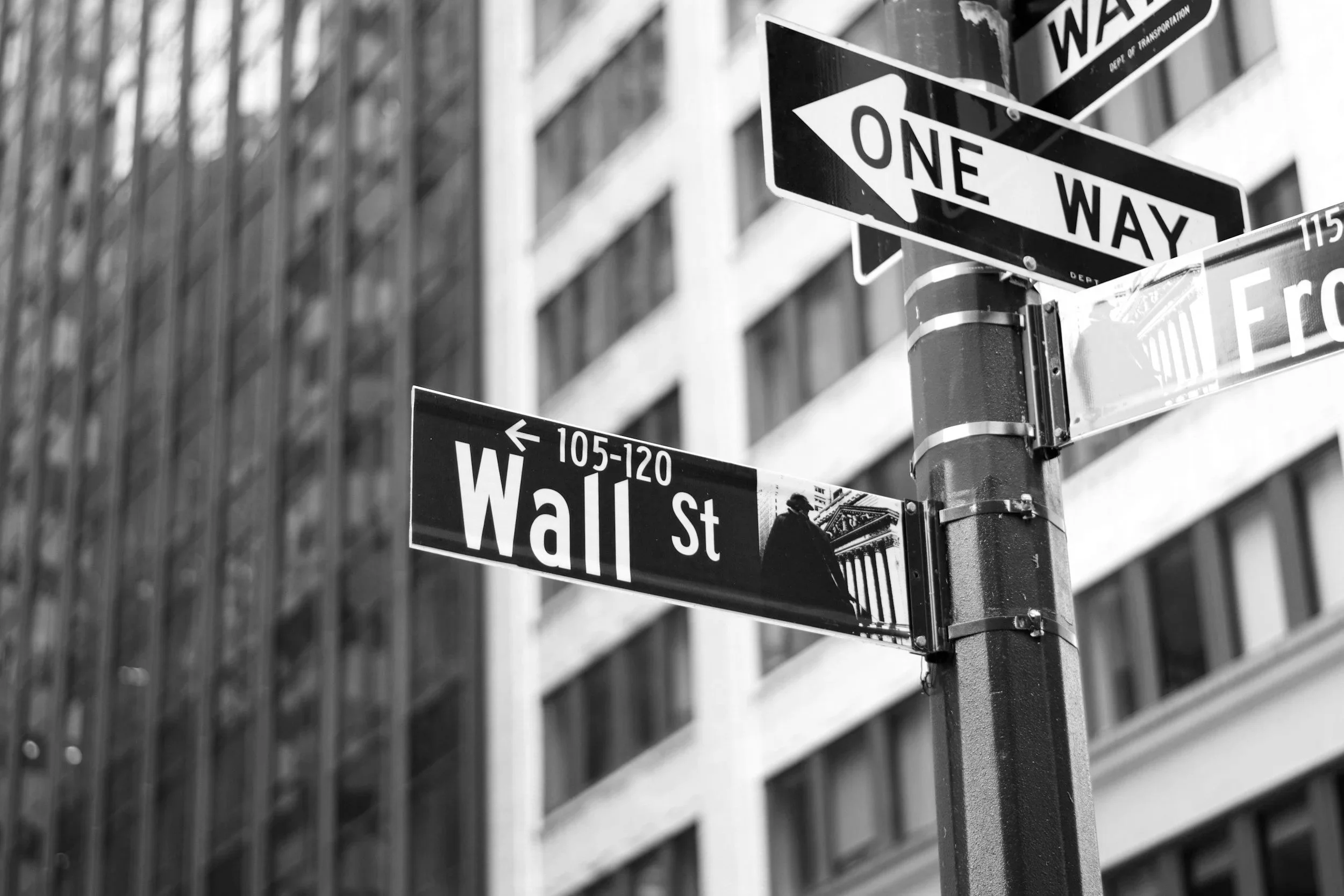 Black and white street corner signs showing 'One Way' with an arrow pointing left, and street name 'Wall St' with a building in the background.